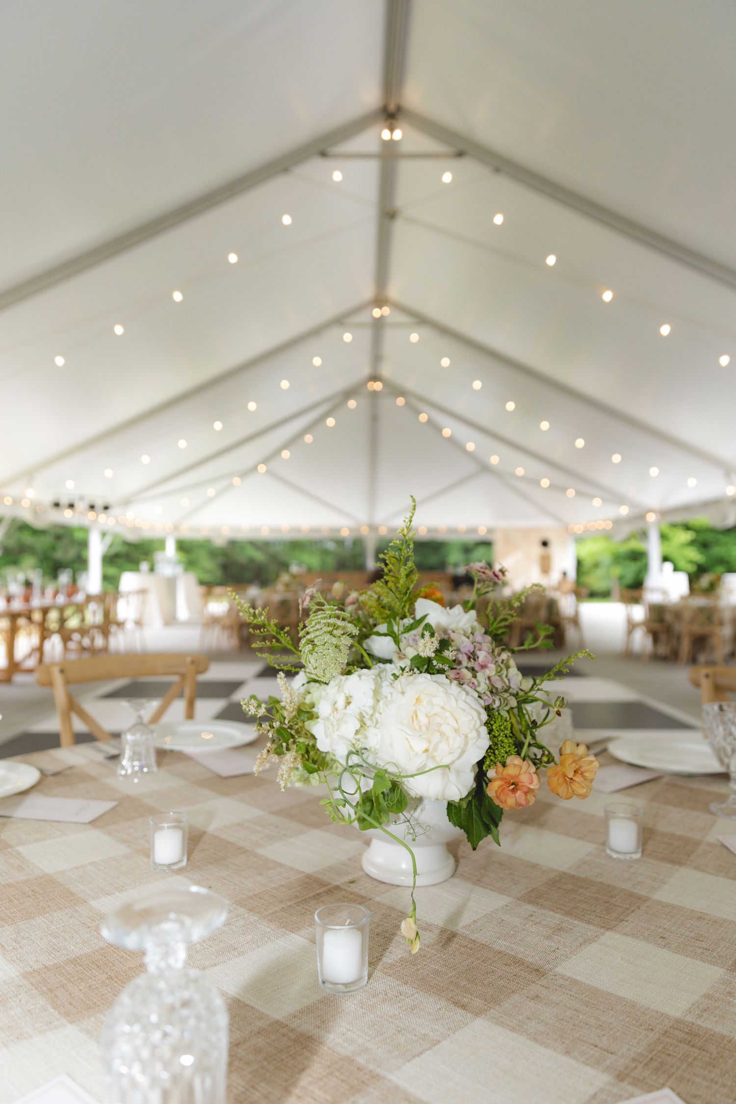 Elegant wedding reception tent with string lights, floral centerpiece of white peonies, and checkered tablecloths.