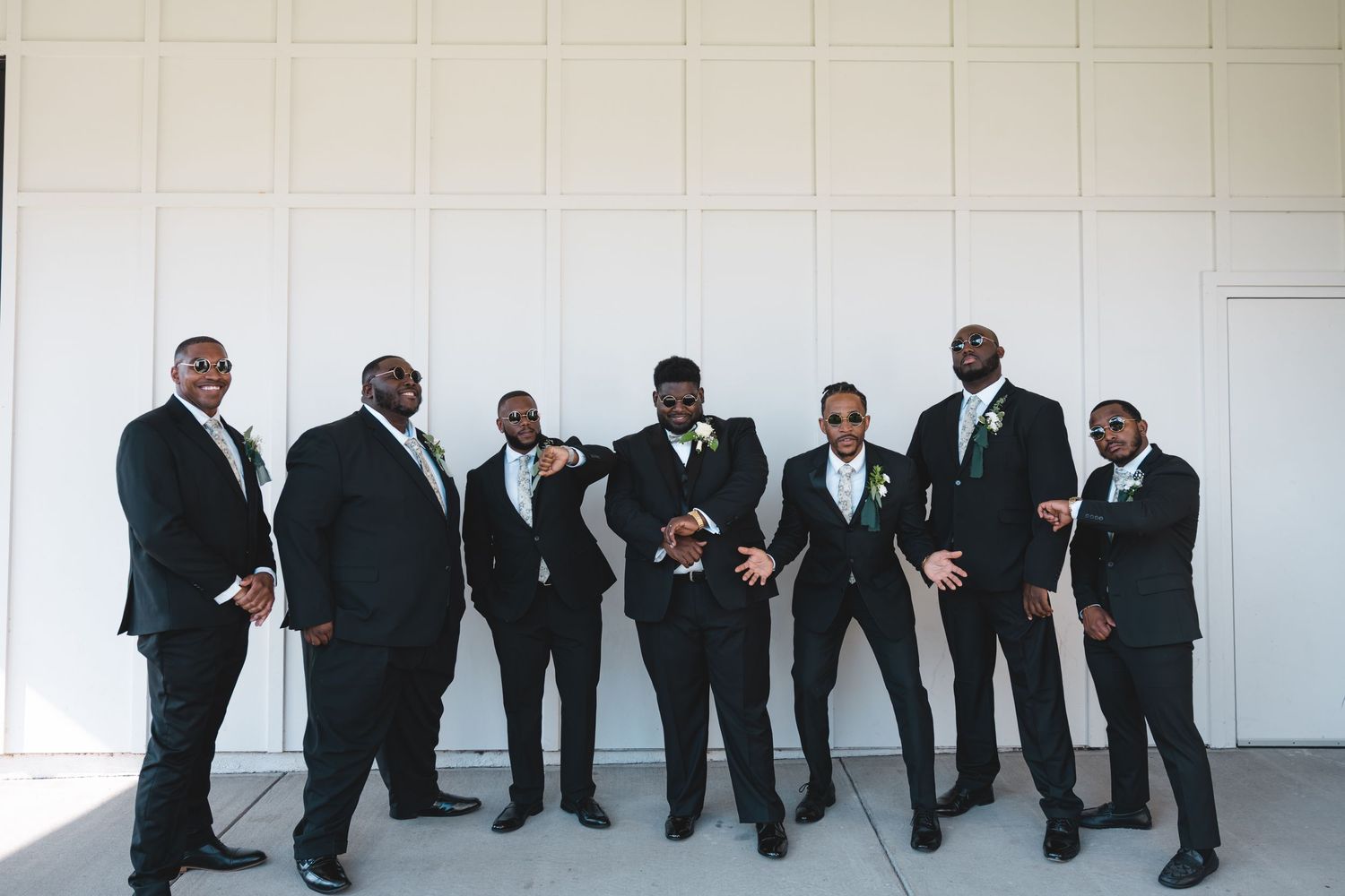 Groom poses with six groomsmen in matching black suits and green ties against a white wall.