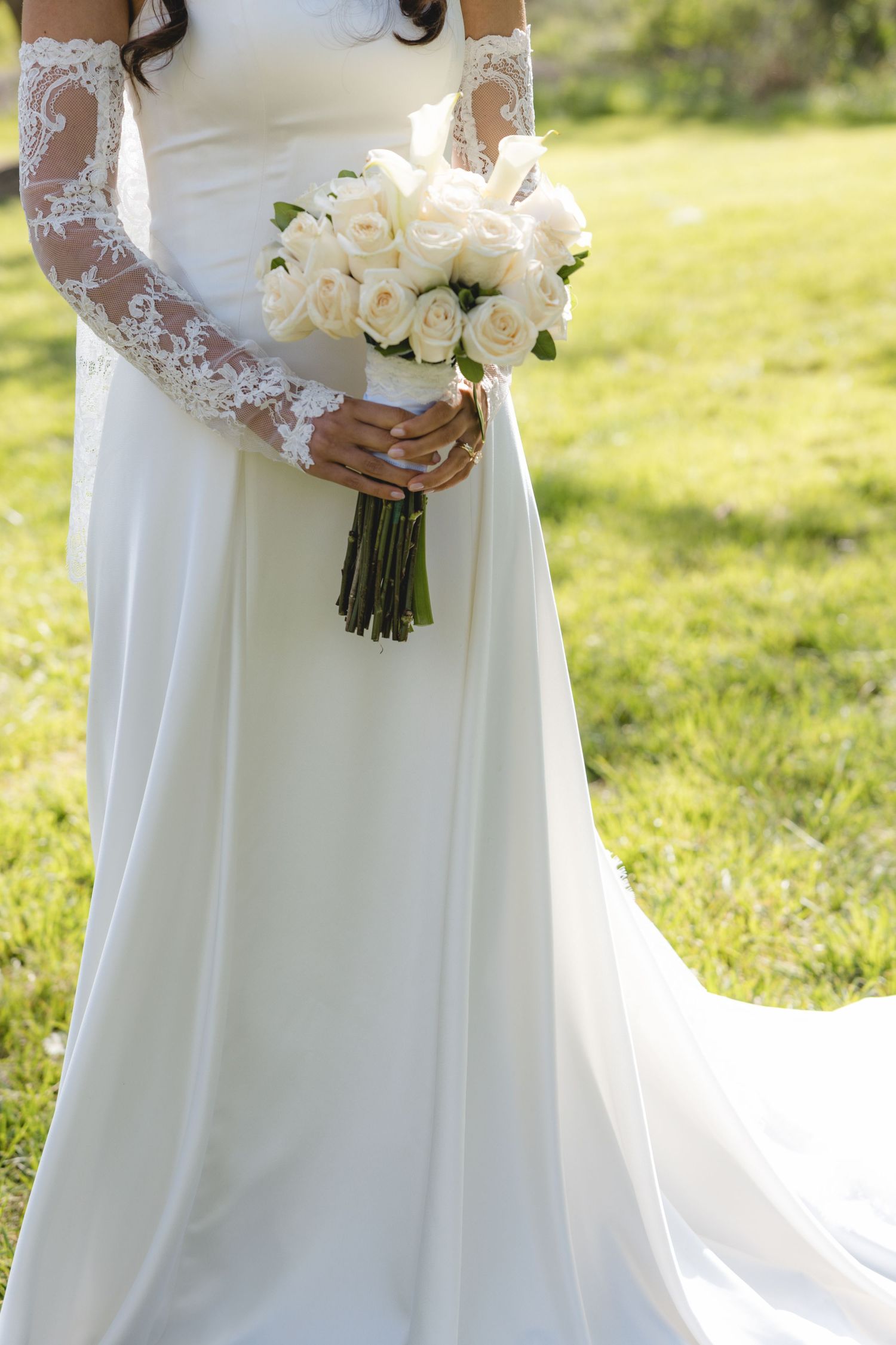 Bride in white lace long-sleeve gown holds classic white rose bouquet on green grass outdoors.