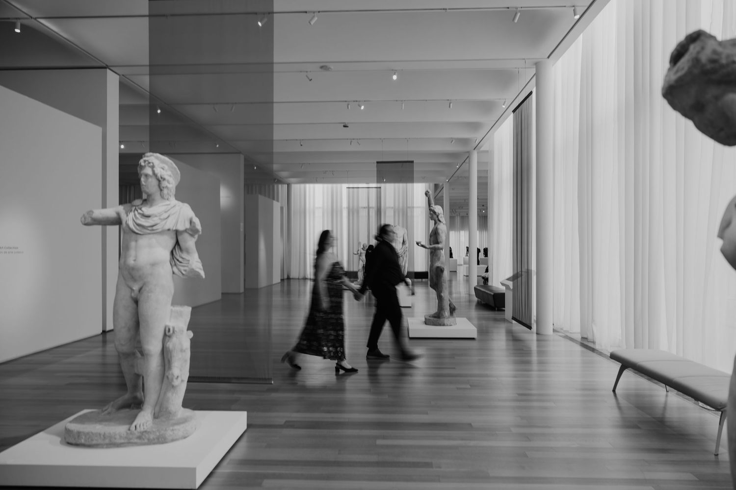 Long minimalist gallery hallway with white walls, sculptures, and visitors creating dramatic shadows.