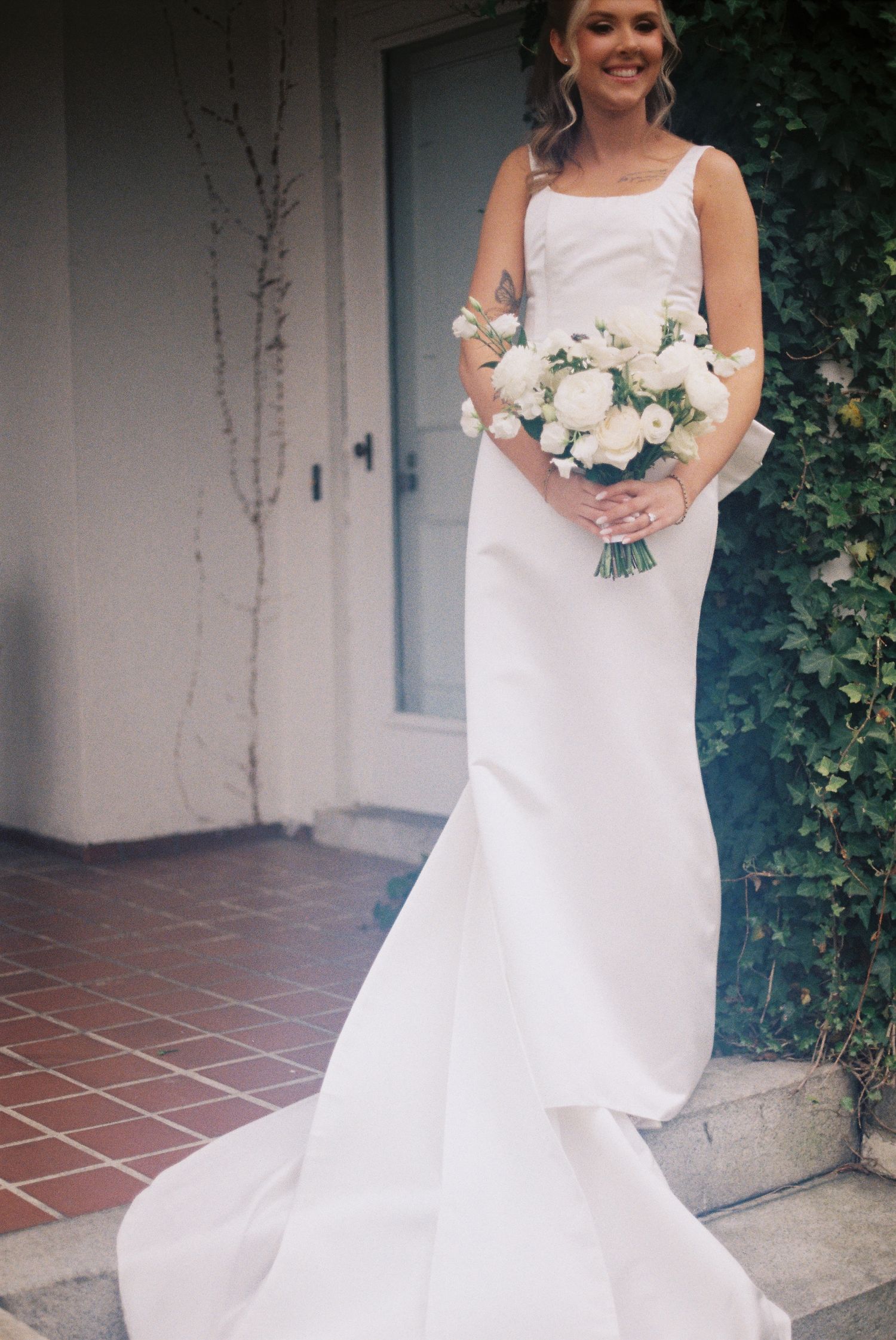 A bride in a white wedding dress stands holding a bouquet of white flowers outside a building.