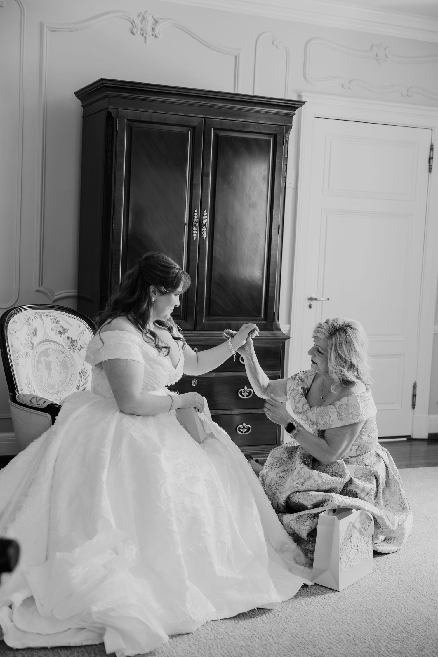 Black and white photo of a bride in a ball gown sitting with a woman helping her with accessories before the wedding.