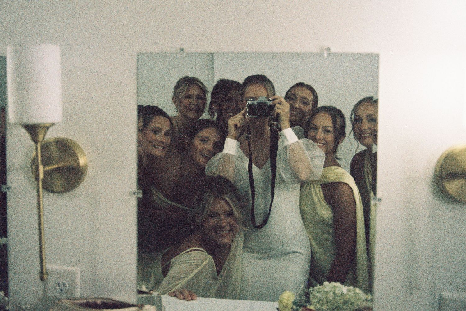 Group of bridesmaids taking a mirror selfie with a camera, smiling together in a bright room with gold sconce lighting.