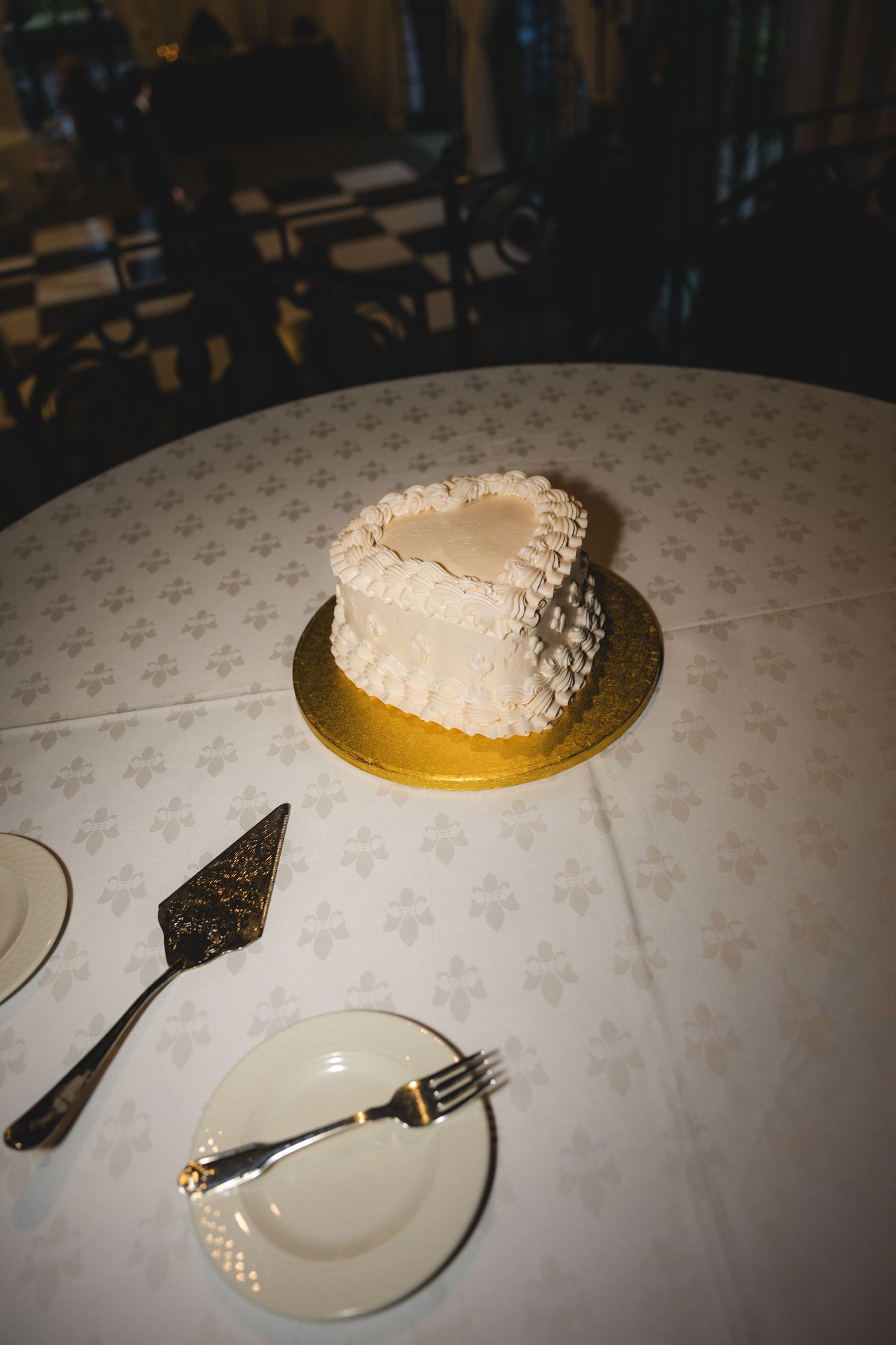 Heart-shaped white wedding cake on gold board with cake server and fork on elegant white tablecloth.