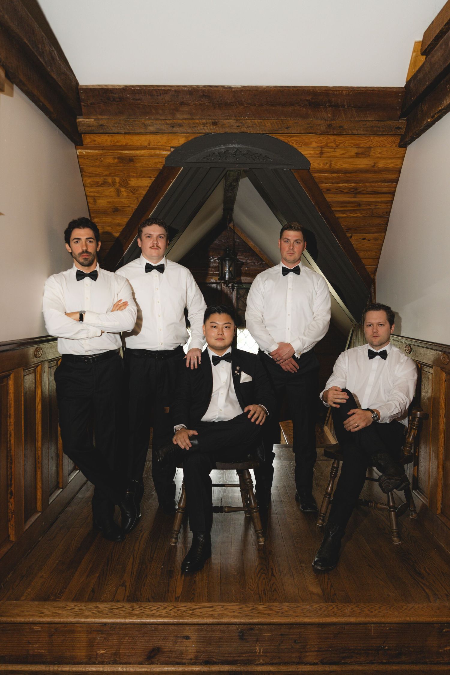 Five groomsmen in white shirts, black pants and bow ties pose on a rustic wooden staircase landing with one seated in front.