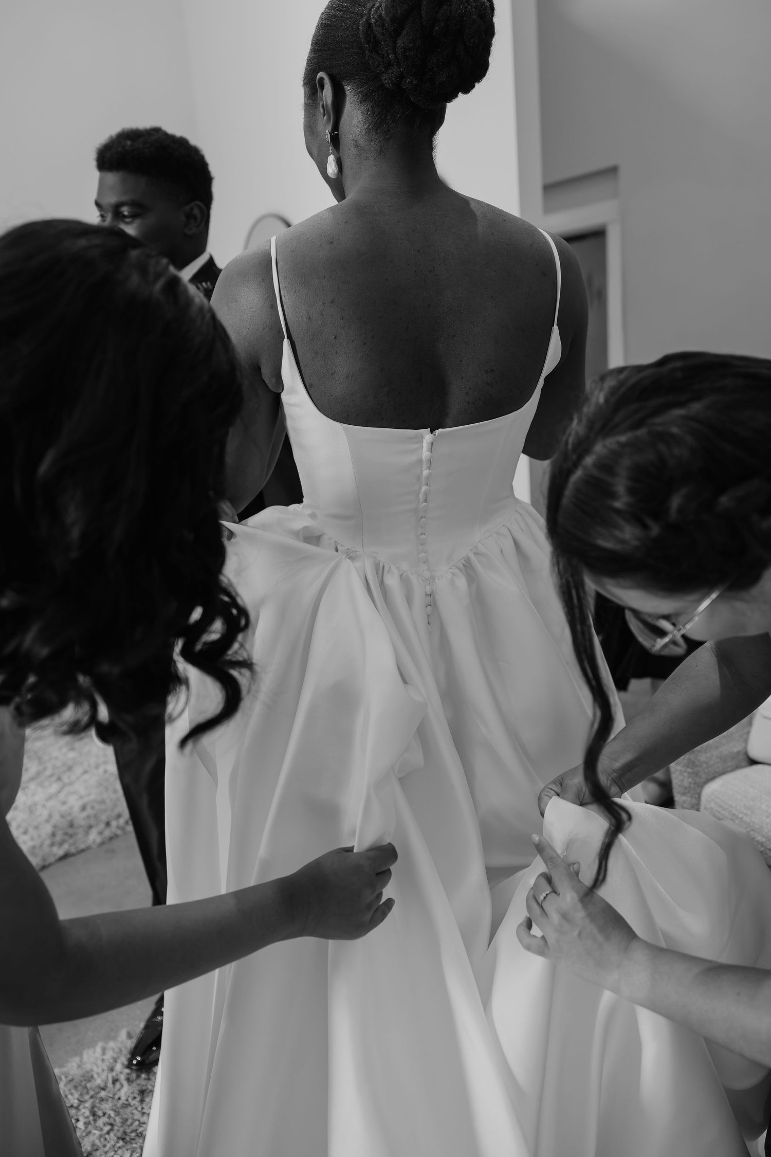 Black and white photo of bride in wedding gown being assisted by bridesmaids buttoning the back of her dress.