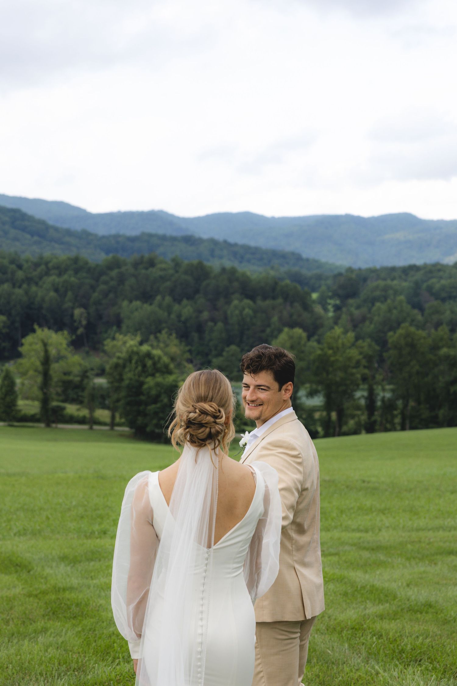 Bride and groom share a romantic moment in a lush green field with mountain views on their wedding day.
