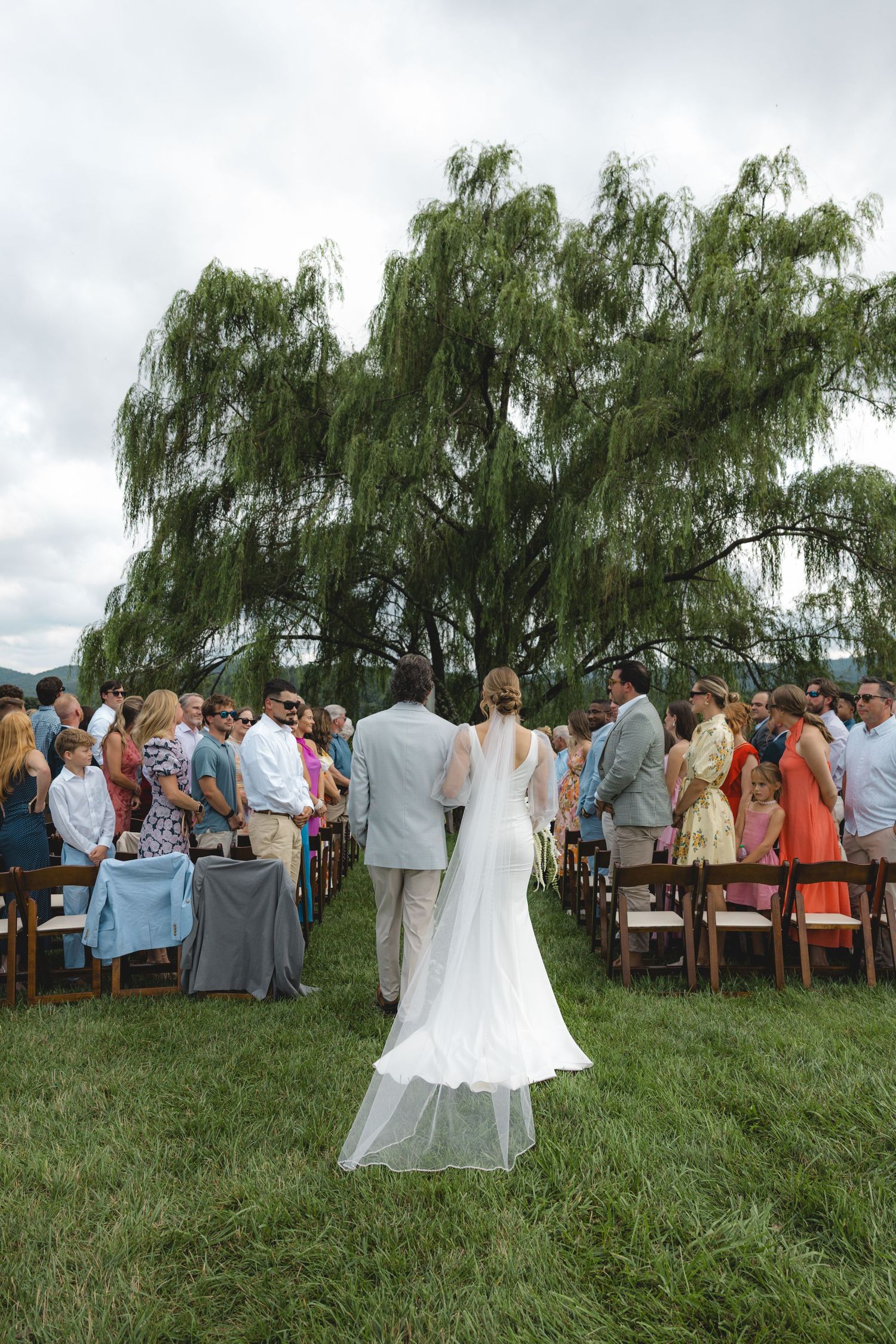 Bride walking down outdoor aisle with escort toward wedding guests seated near weeping willow tree.