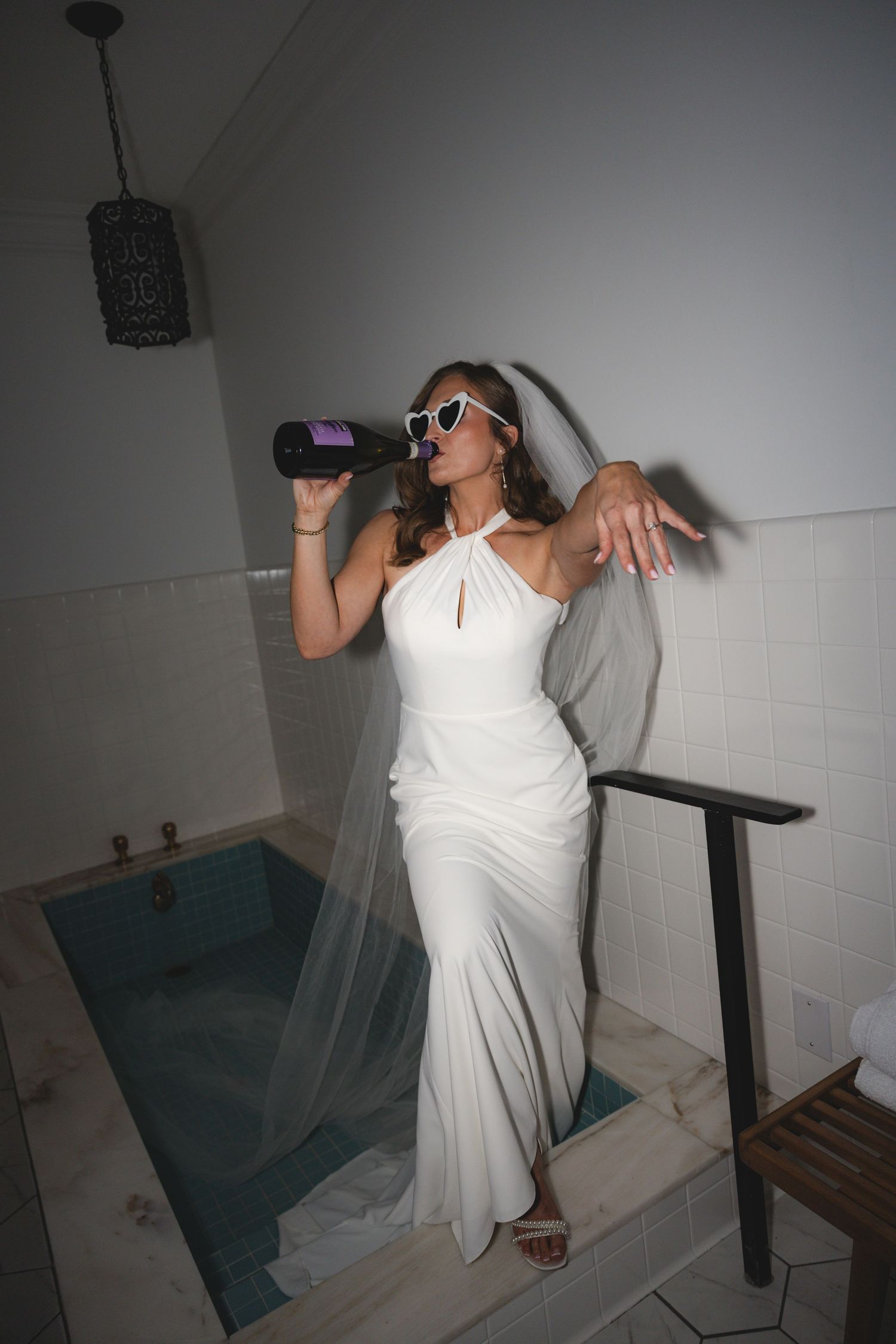 Bride in white halter gown and sunglasses drinking champagne by an indoor pool, wearing a veil.