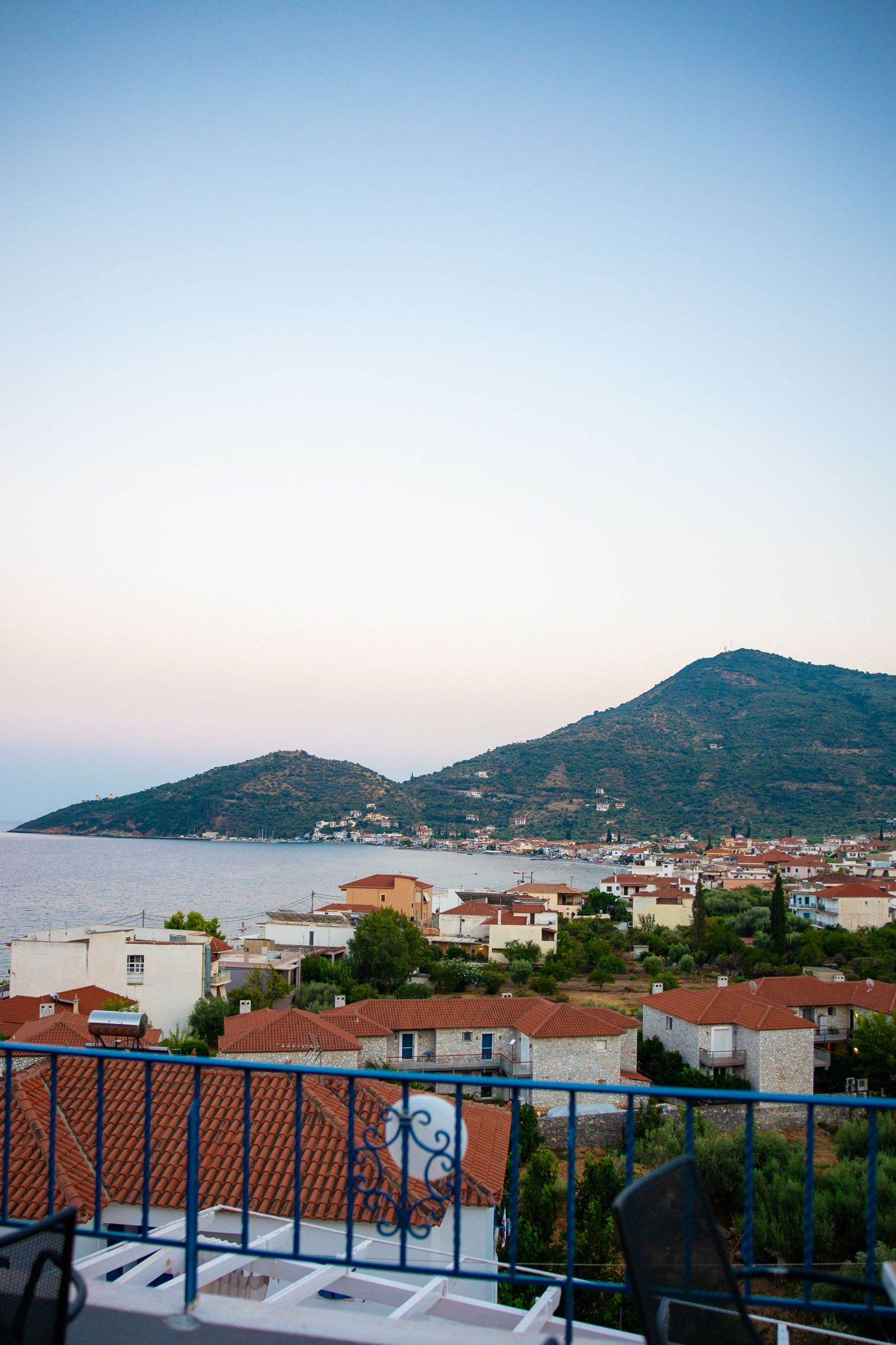 Scenic coastal town view from balcony at dusk, with red-roofed buildings, blue sea, and green mountain backdrop.