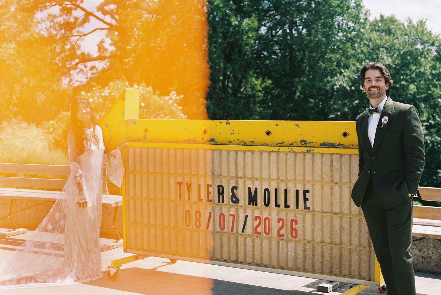 Couple Tyler and Mollie pose by a yellow sign marking their wedding date 08/07/2026 outdoors.