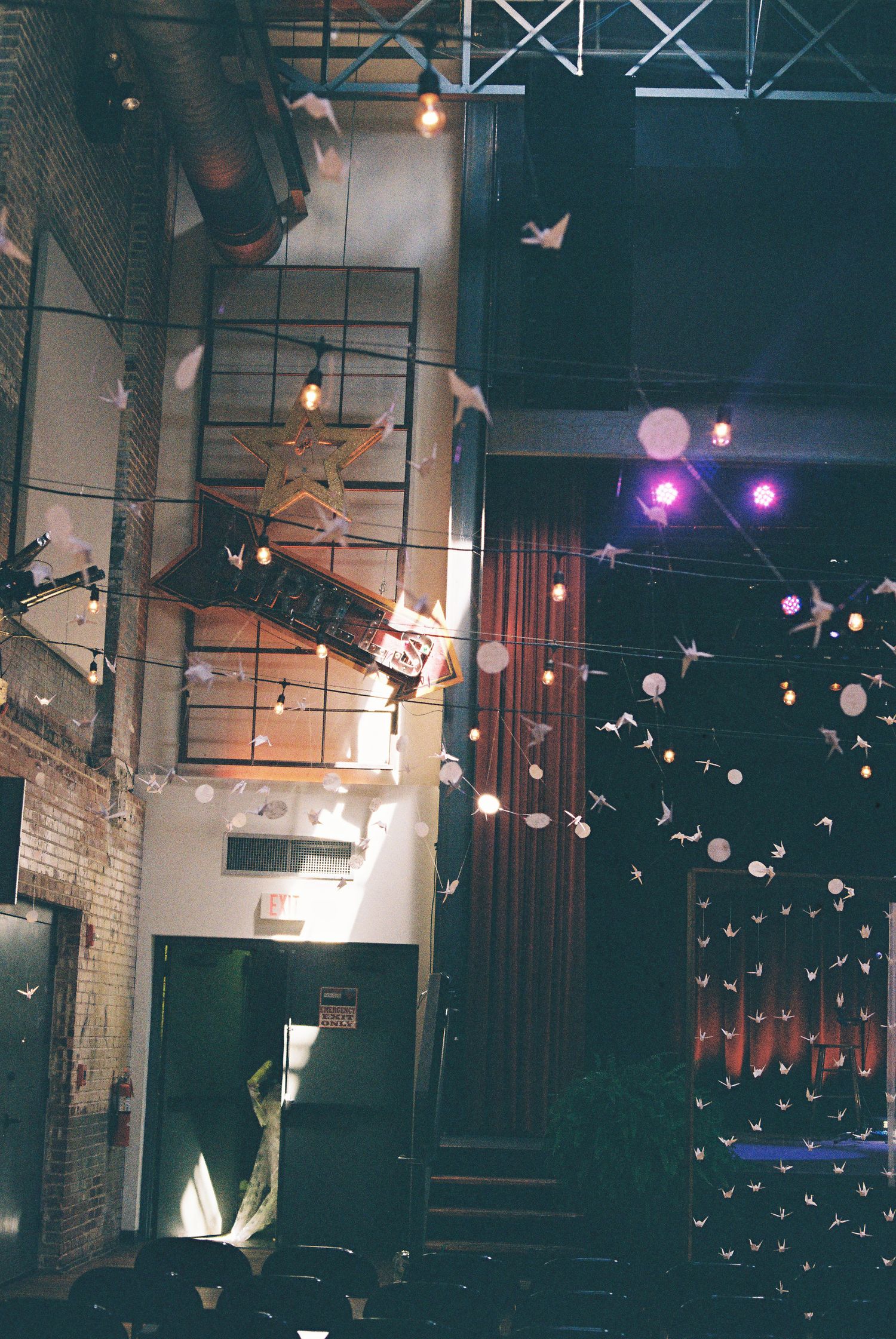 Industrial venue interior with string lights, paper cranes, and stage lighting creating a whimsical atmosphere.