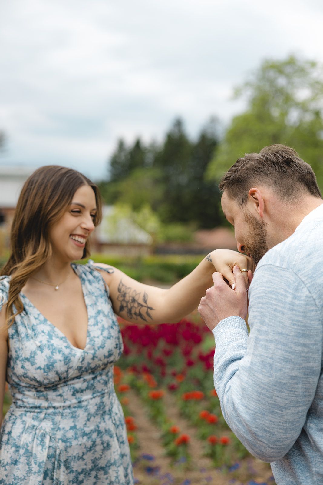 A man kisses a smiling woman's hand in a colorful tulip garden, as she shows off her tattoo in a floral dress.