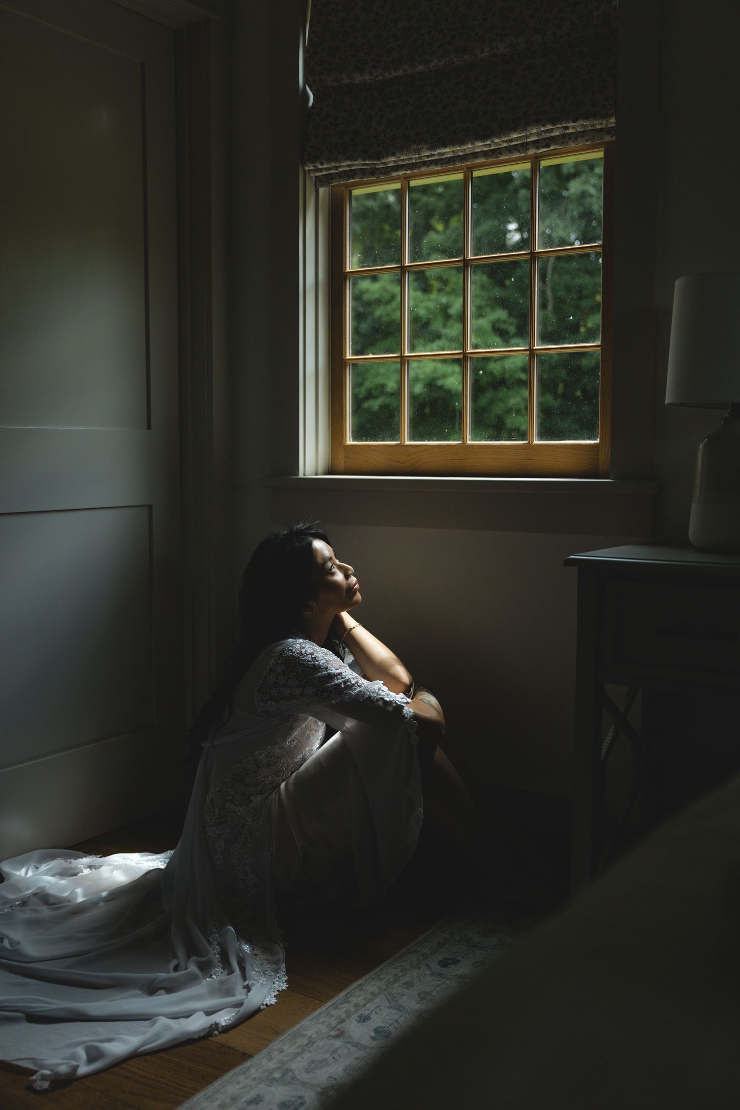 Woman in wedding dress sitting on floor by sunlit window in dark room, contemplative mood.