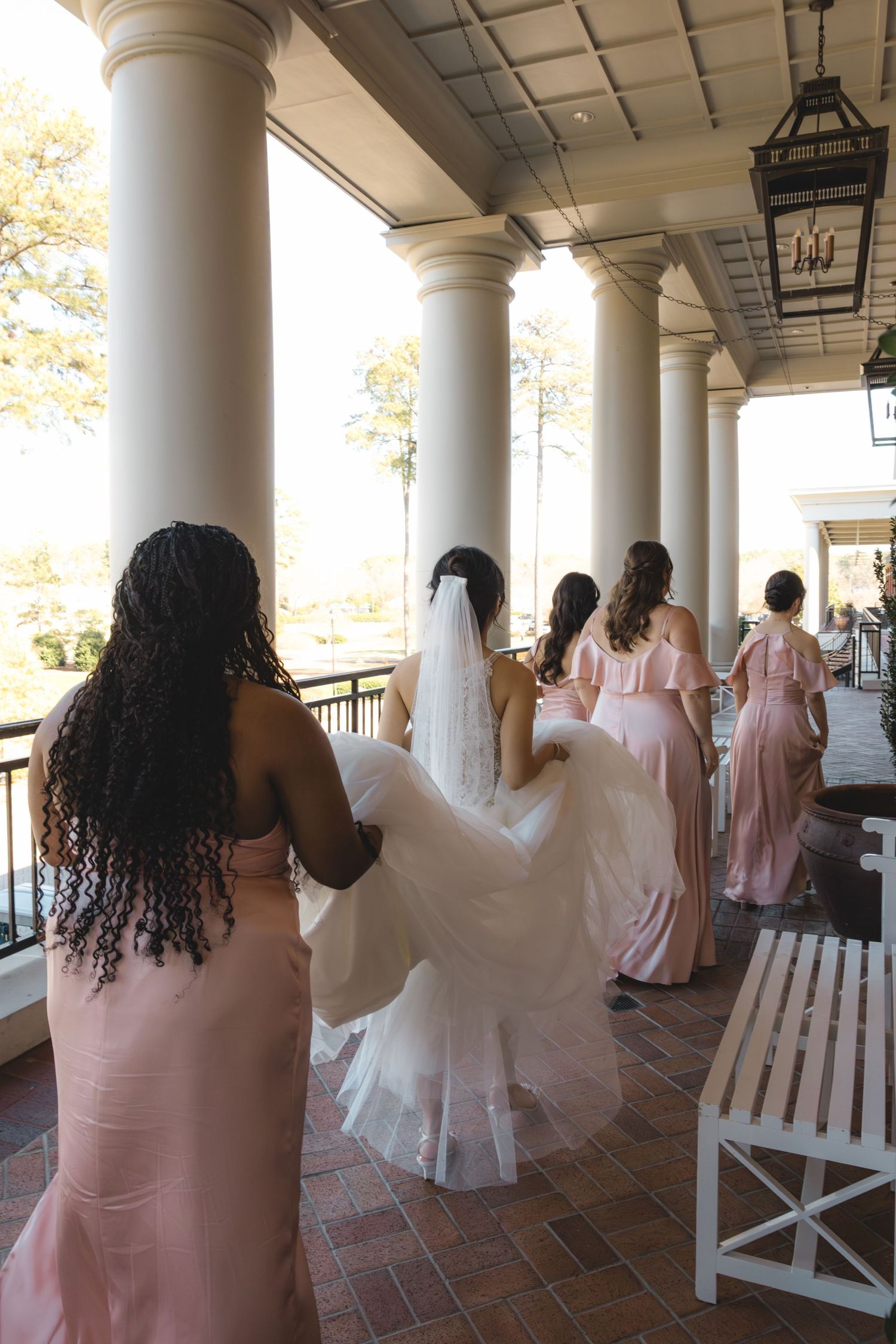 Diverse bridesmaids in blush pink gowns escort veiled bride along columned outdoor venue portico on wedding day.