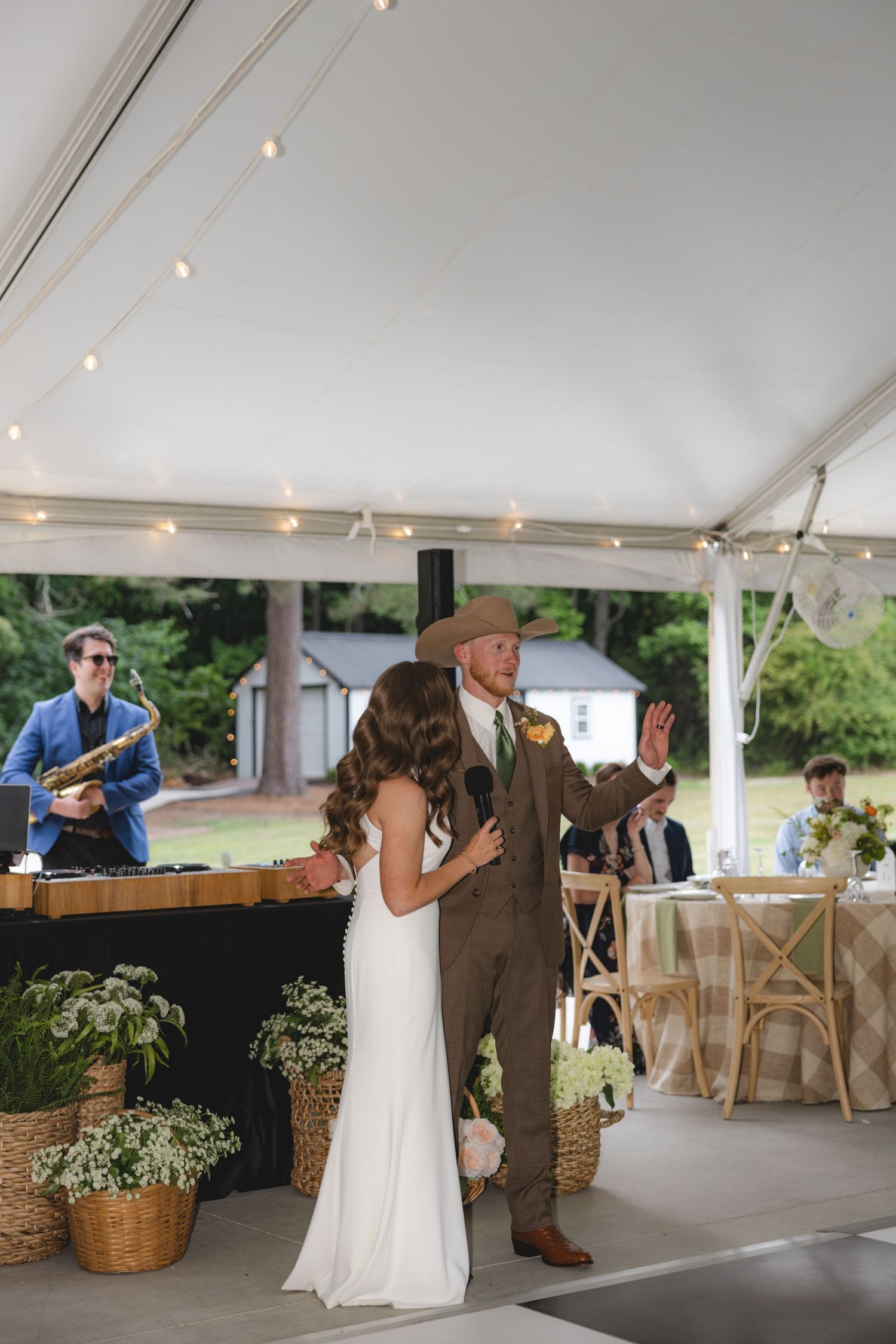 Groom in cowboy hat addresses wedding guests with microphone alongside bride at tented reception.
