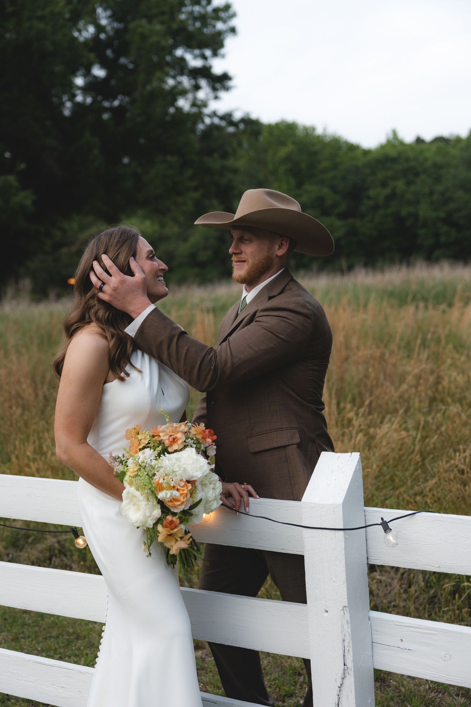 Bride in white gown holding floral bouquet with groom in brown suit and cowboy hat by white fence in field.