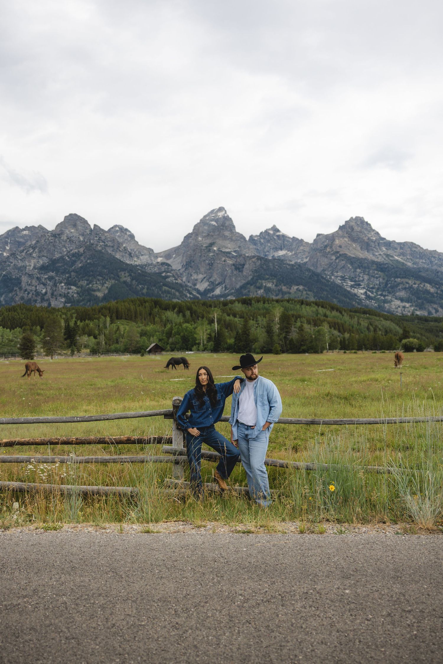 Two people in denim lean on a wooden fence with Grand Teton mountains, green fields, and horses in the background.