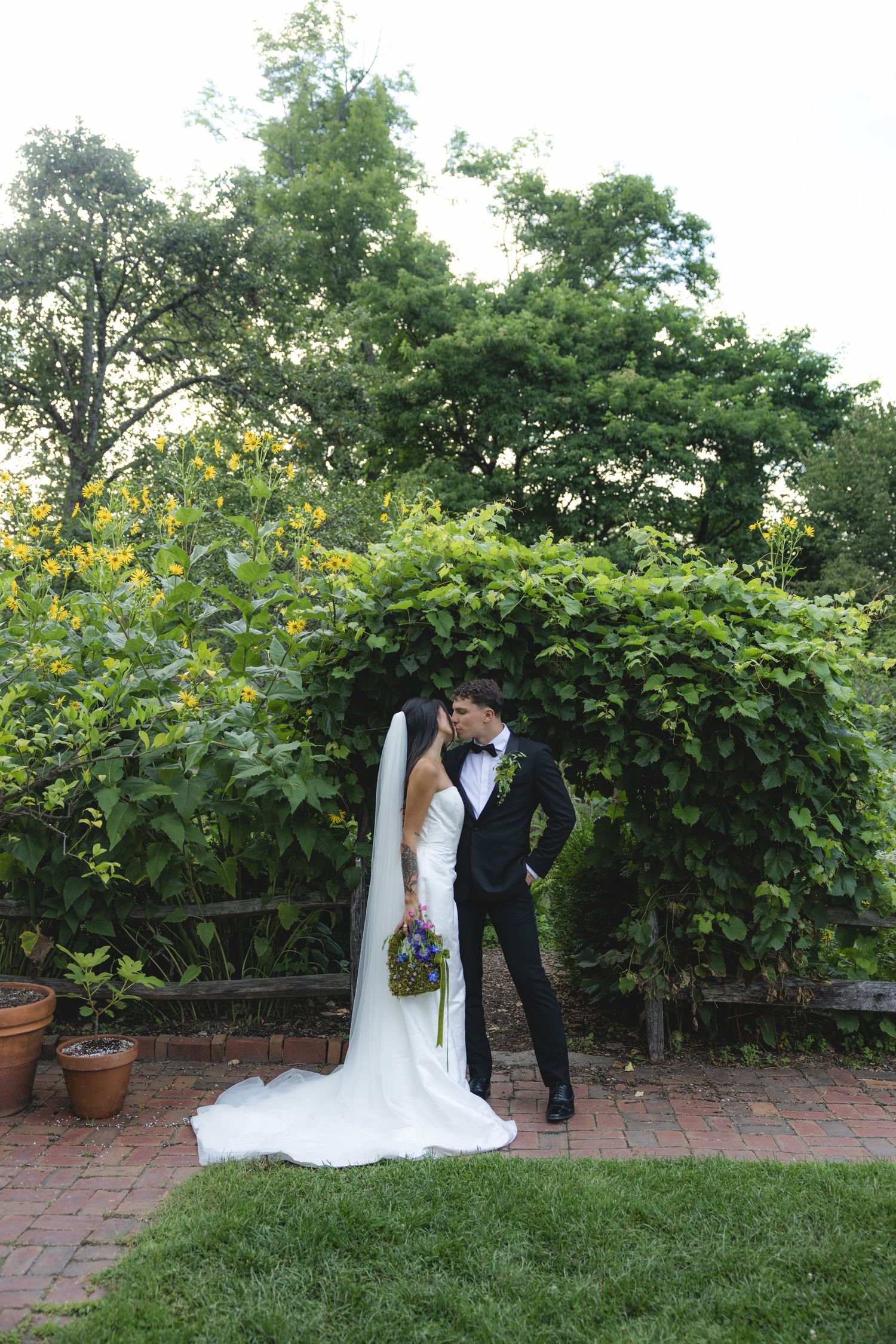 Bride and groom share a romantic kiss in a lush garden setting, bride in white gown with veil holding bouquet.