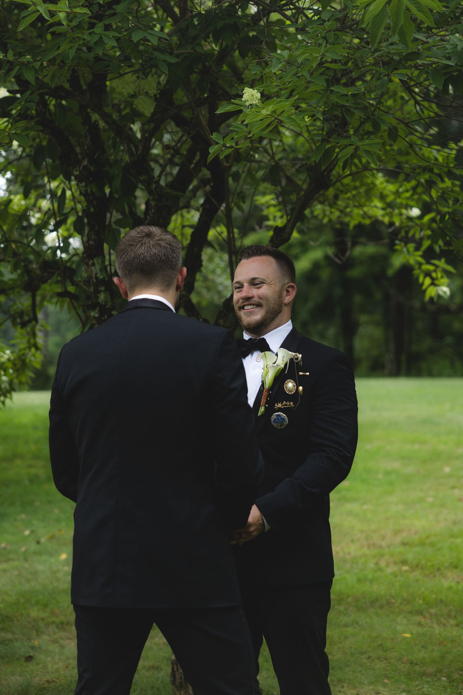 Two grooms in black tuxedos share a joyful moment outdoors during their wedding ceremony.