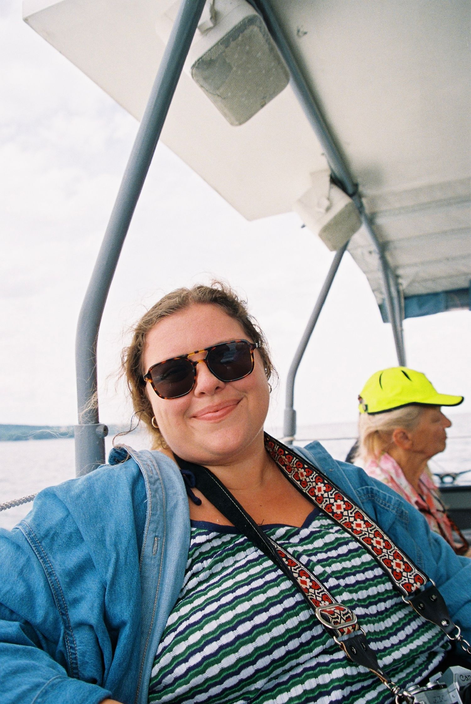 Woman in sunglasses and striped shirt smiling on a boat, wearing a floral crossbody bag on a cloudy day.