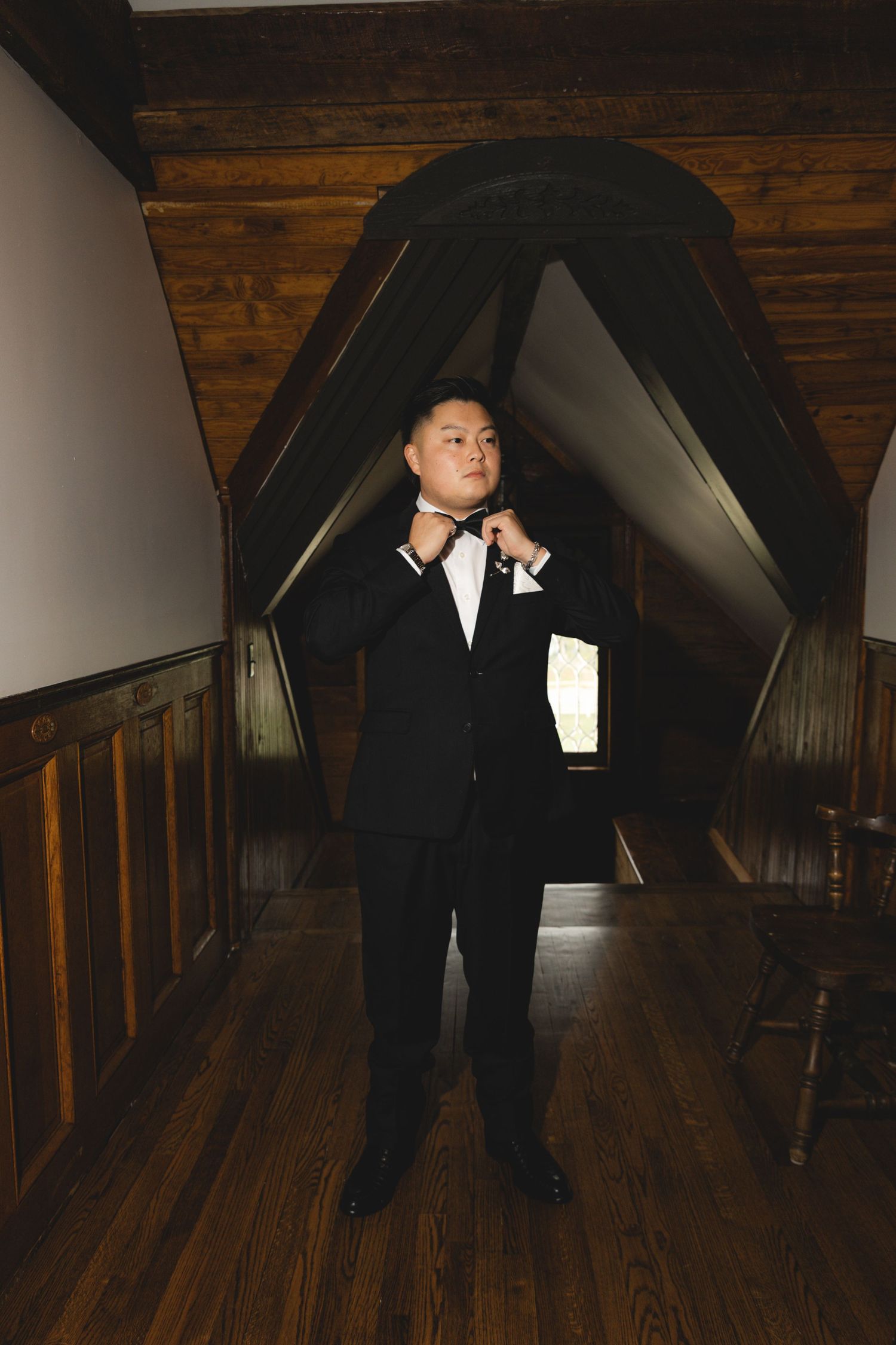 Groom in a black tuxedo adjusts his bow tie in a rustic attic room with wood-paneled walls and hardwood floors.