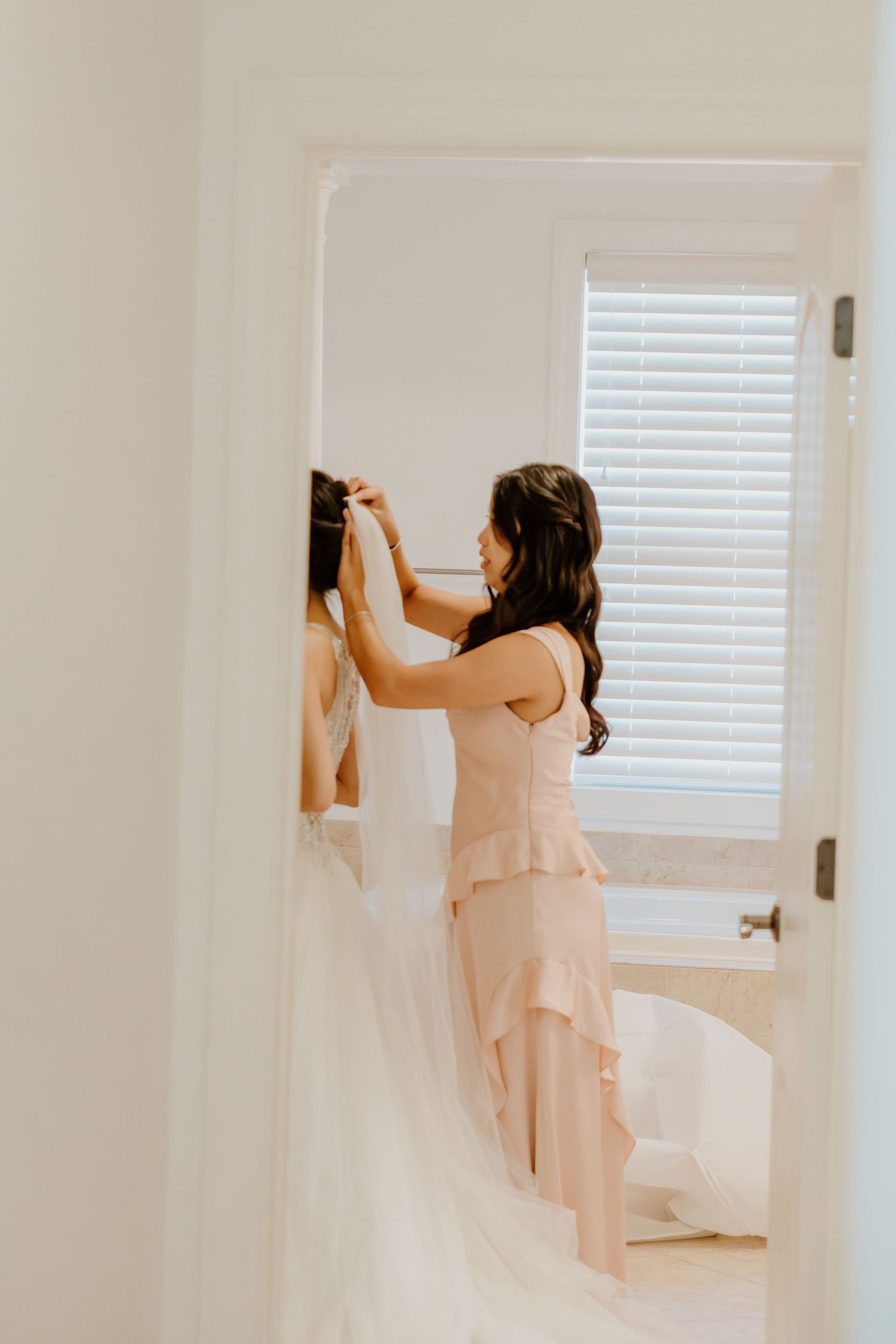 Bridesmaid in blush ruffled gown helps bride with wedding dress details in a bright white bathroom doorway.