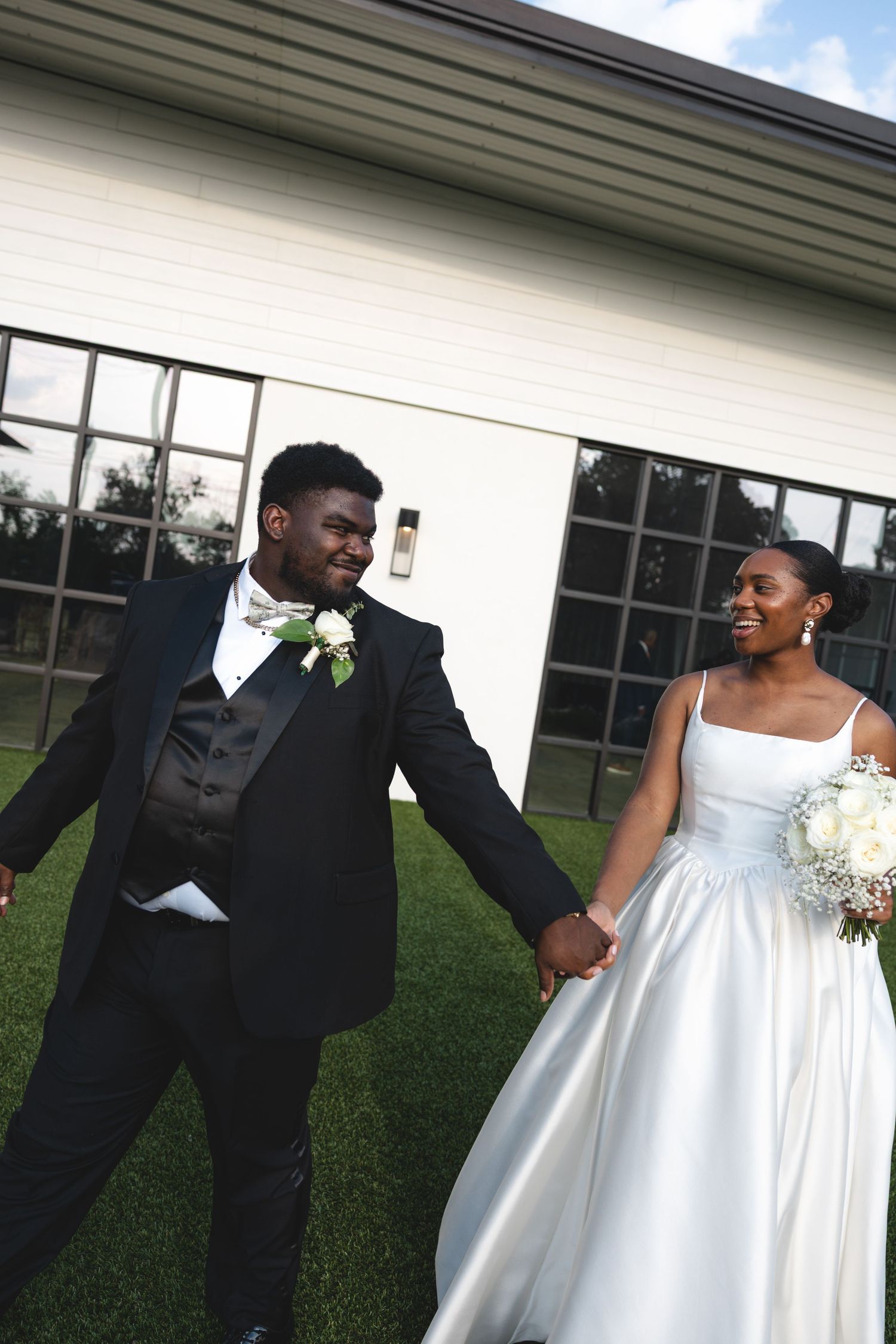 Newlyweds hold hands and walk smiling outside wedding venue.