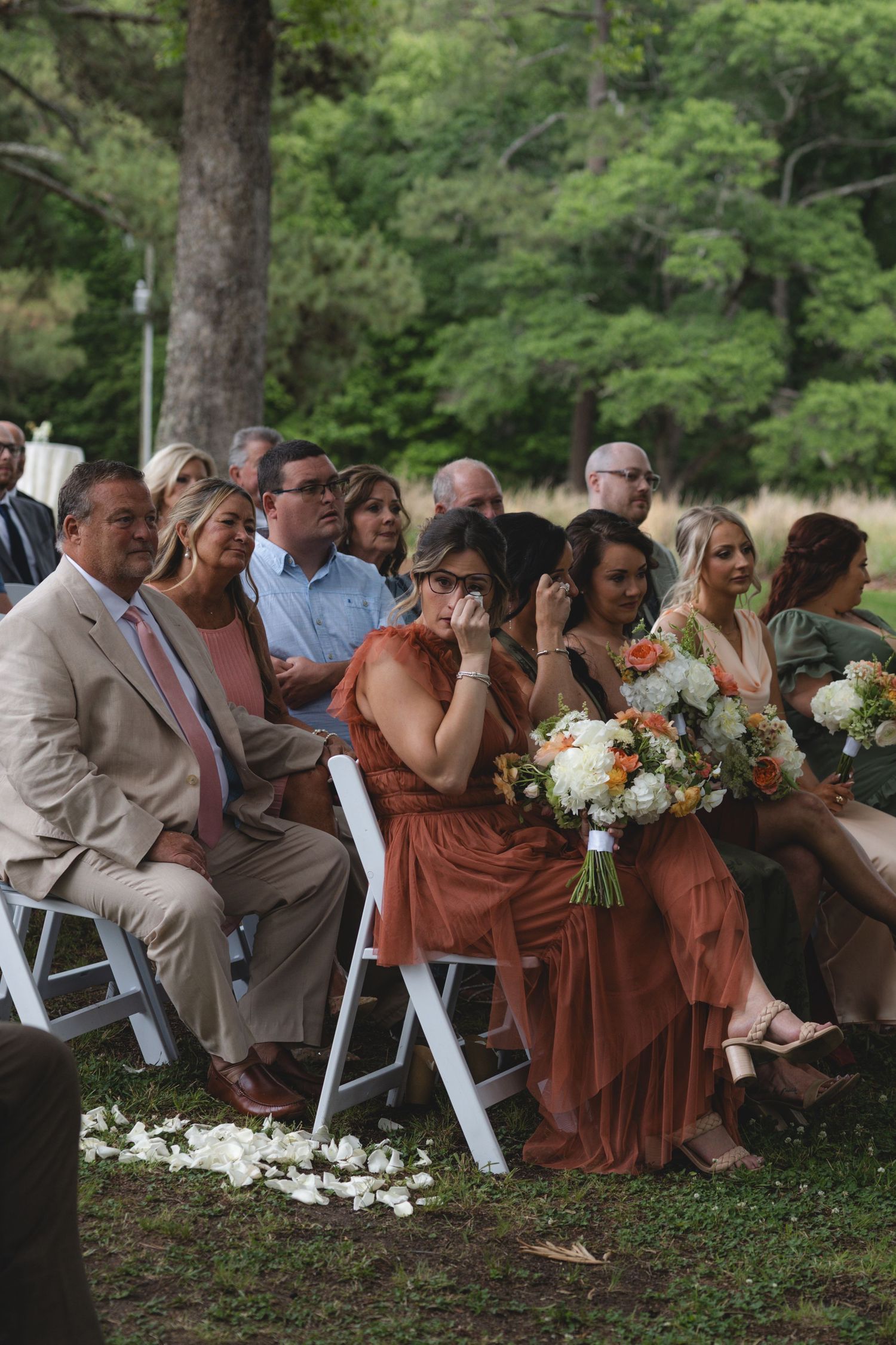 Emotional wedding guests seated in white chairs watch outdoor ceremony, bridesmaid holds orange and white bouquet.