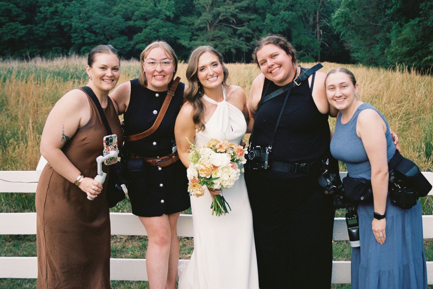A bride in white holding a floral bouquet poses with four friends outdoors near a wooden fence.