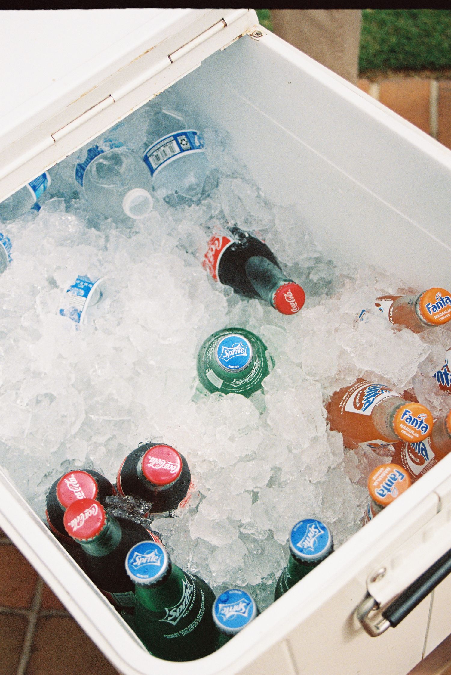 Assorted soda and beer bottles chilling in ice inside a white cooler, including Fanta and Sprite.