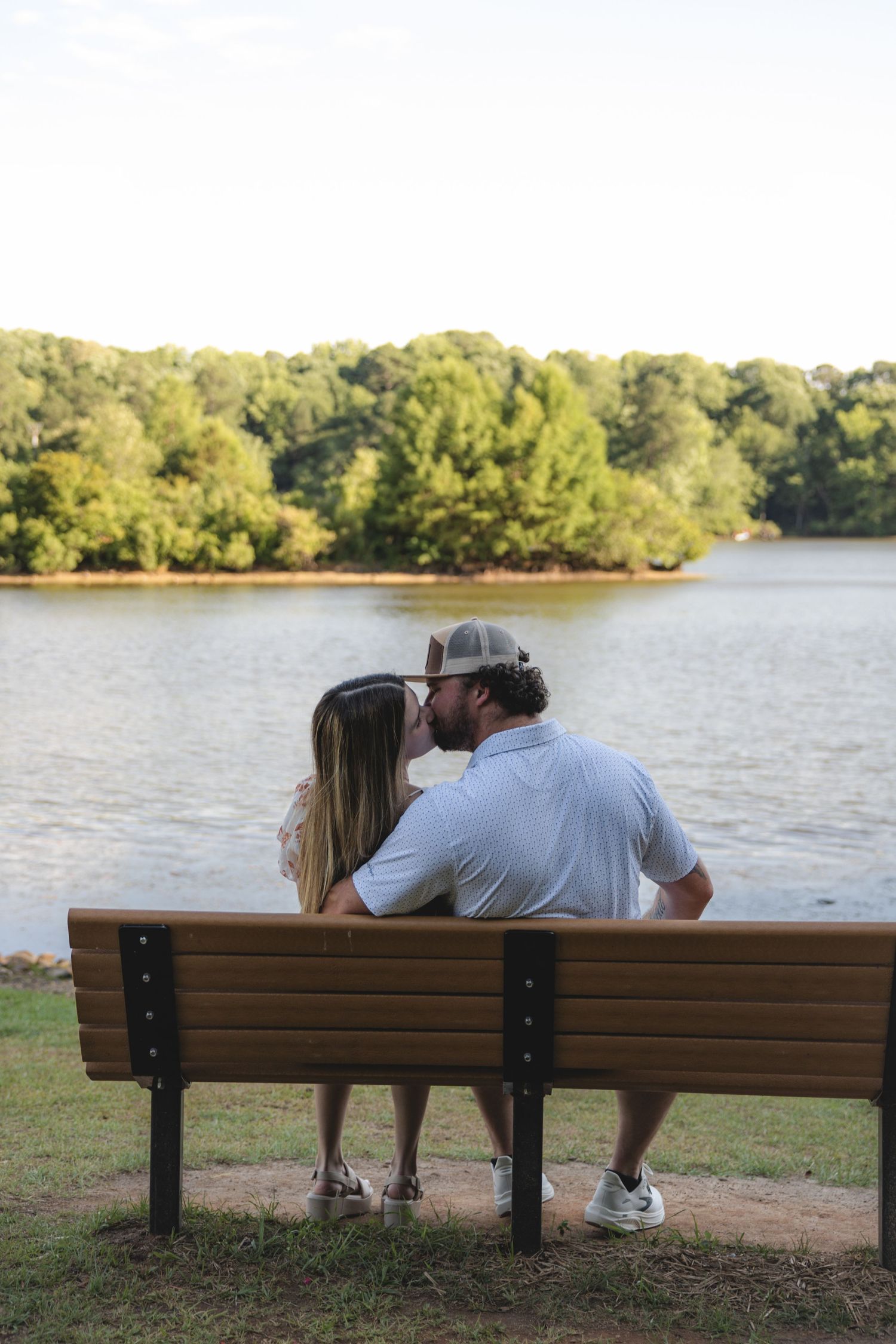 Couple sharing a kiss on a park bench overlooking a serene lake surrounded by lush green trees.