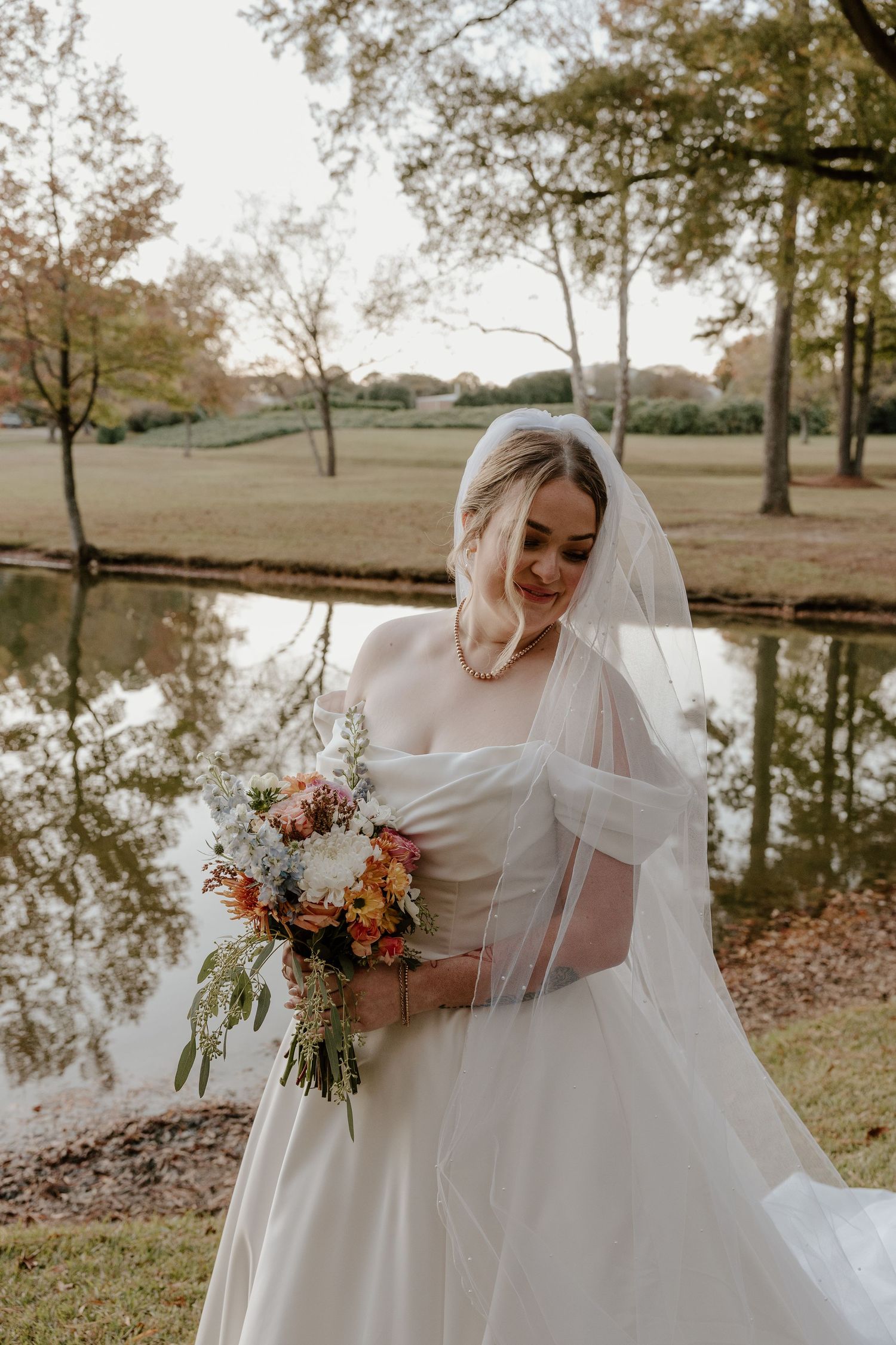 Bride in white off-shoulder gown and veil holds colorful bouquet by a reflective pond in autumn.