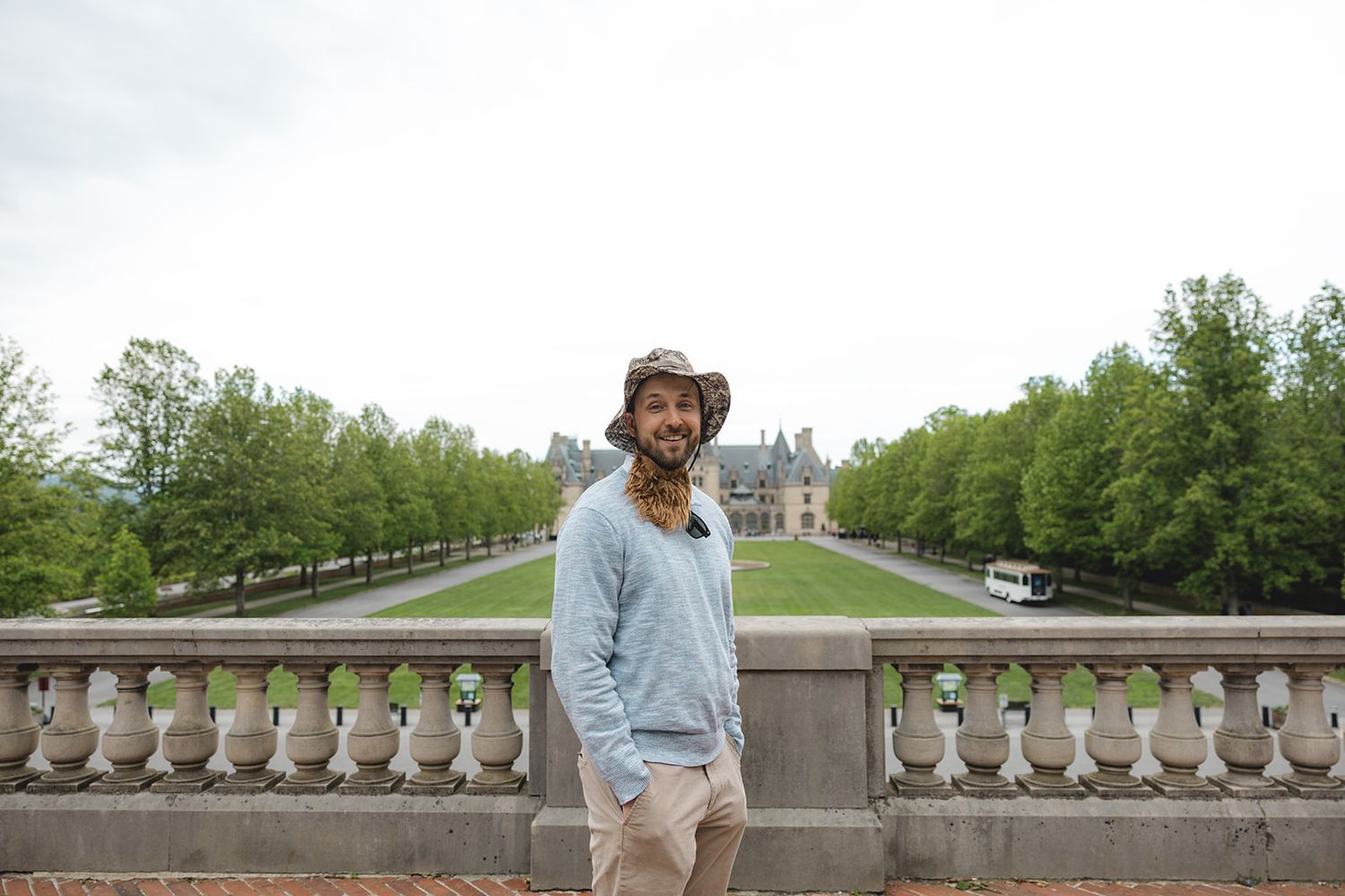 Man in hat smiling on stone balcony with lush green trees and grand estate in background.