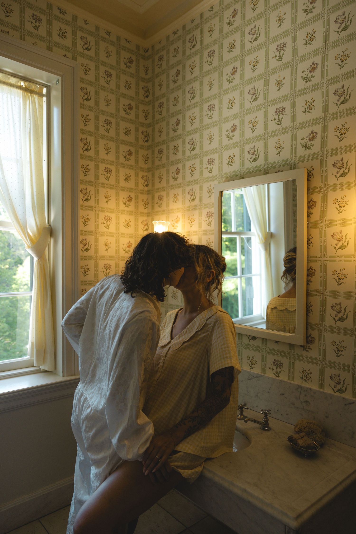 Two people sharing a romantic kiss in a vintage bathroom with floral wallpaper, natural window light, and a wall mirror.