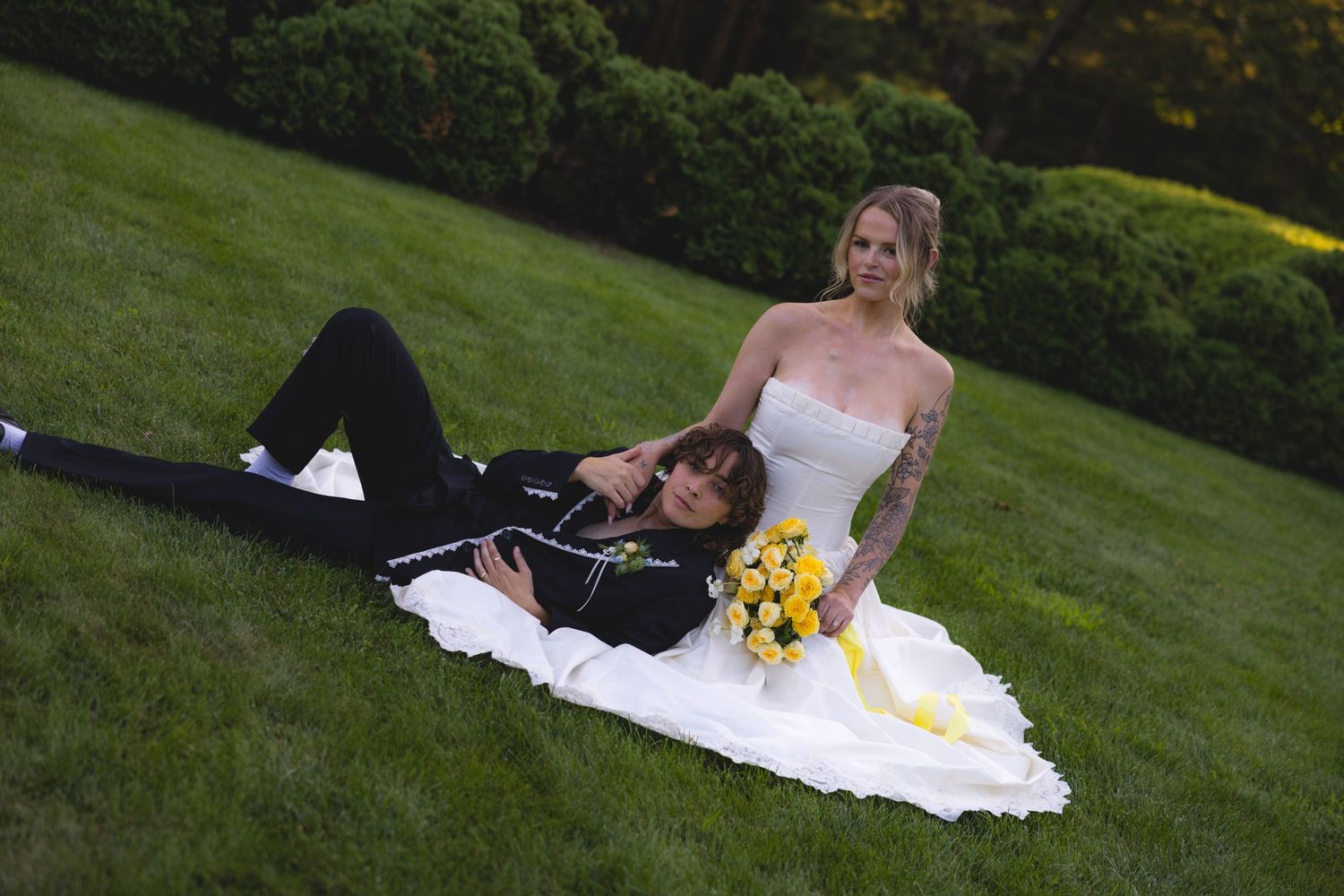 Bride in white strapless gown with yellow bouquet poses on grass with groom in black suit at outdoor wedding.