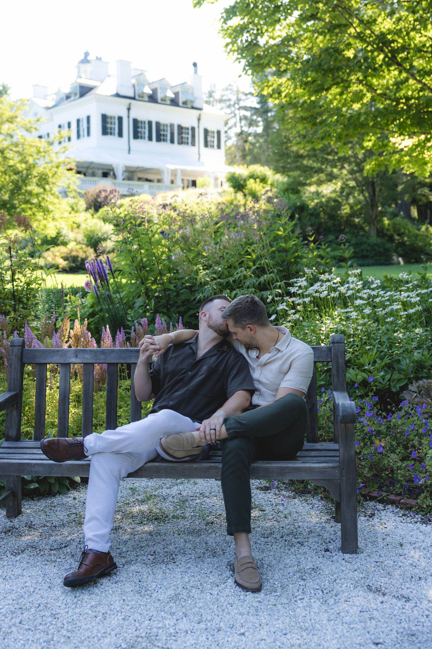 Two people share a romantic moment on a wooden bench in a lush garden with a white house backdrop.