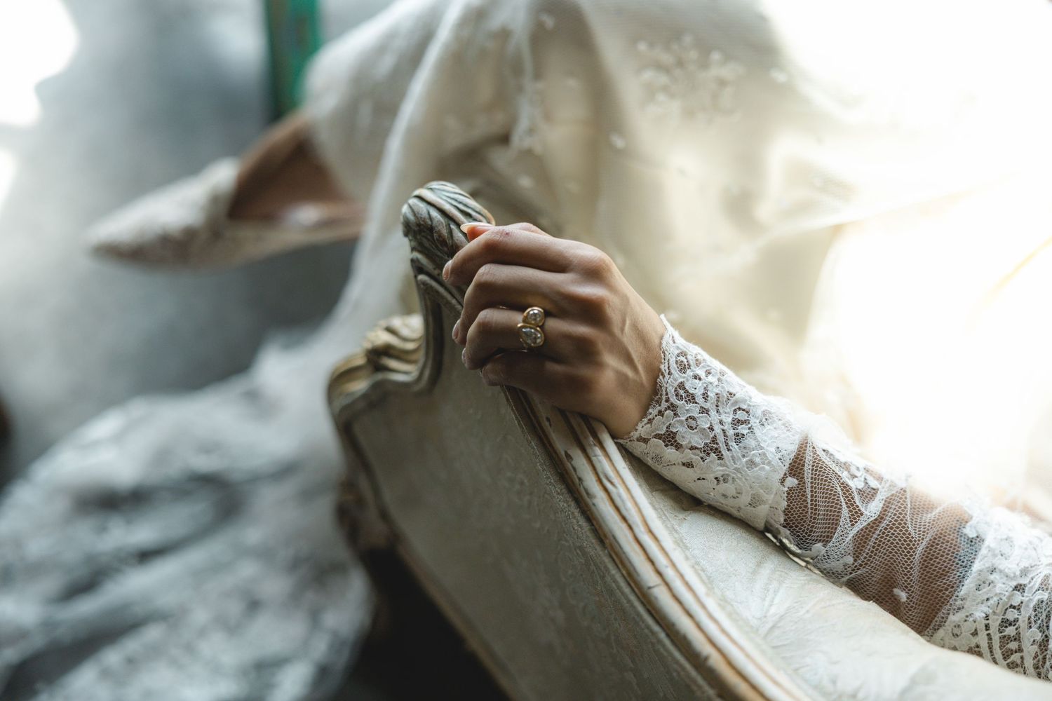 Close-up of a bride's lace-gloved hand resting on a vintage chair, adorned with an elegant ring.