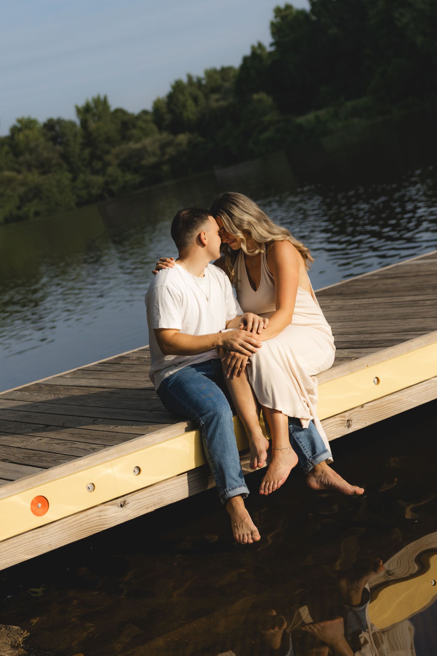 A couple sits barefoot on a wooden dock by a lake, sharing a romantic moment surrounded by lush green trees.