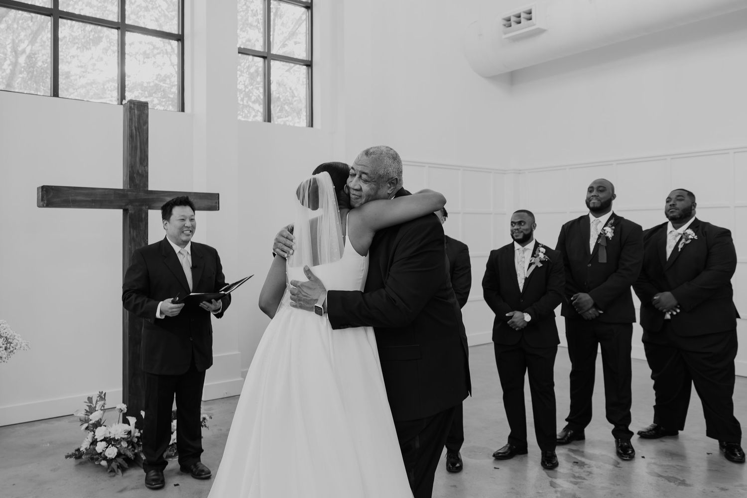 Black and white wedding ceremony photo of bride and groom embracing at altar with groomsmen and officiant.