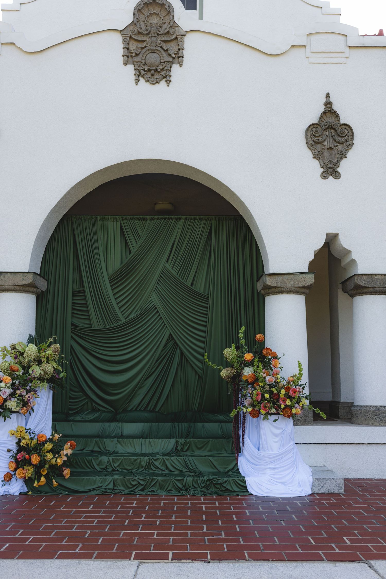 Elegant arched doorway with green draped curtains and floral arrangements on a white building.
