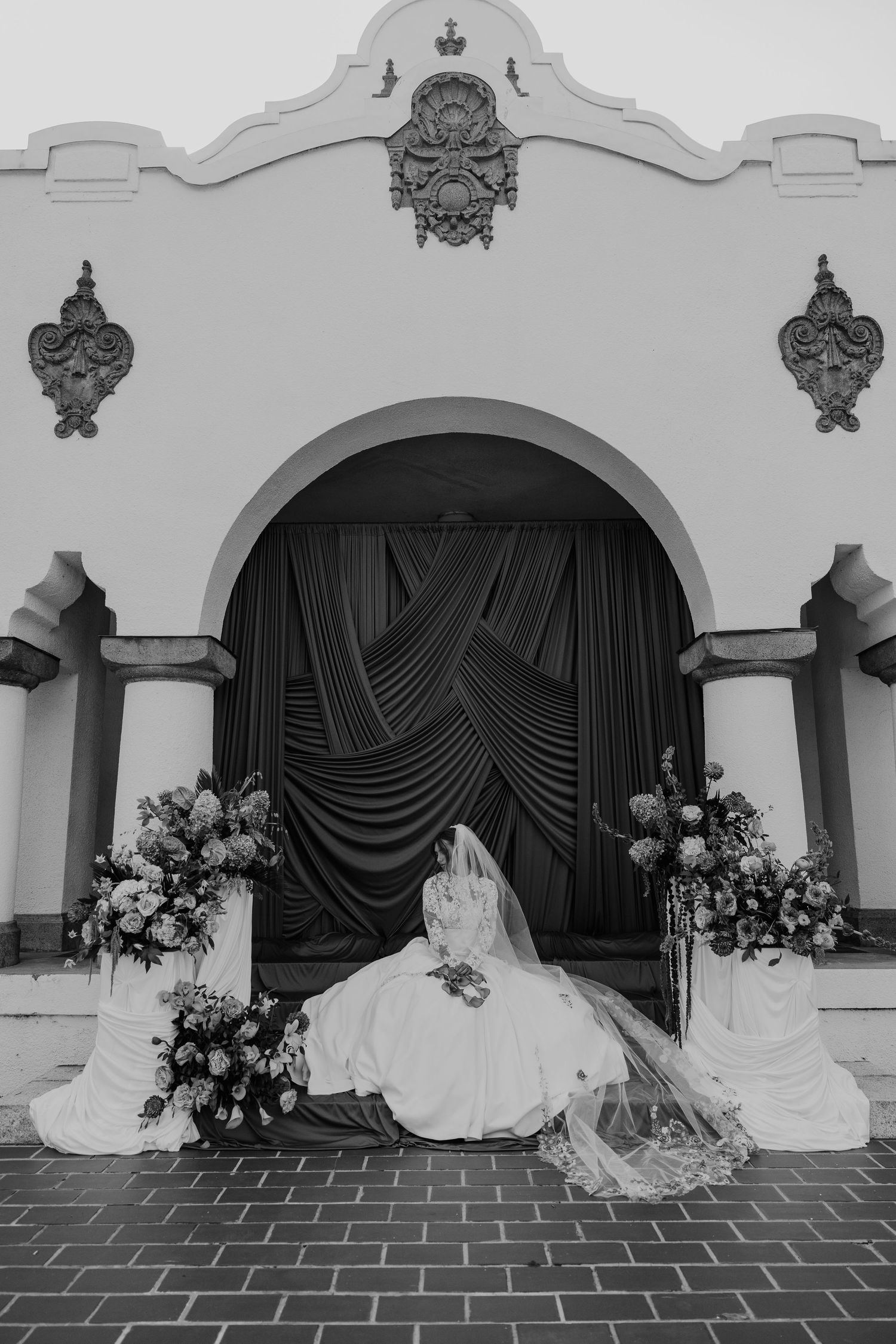Black and white photo of a bride in a flowing gown and veil seated before an arched doorway with floral arrangements.