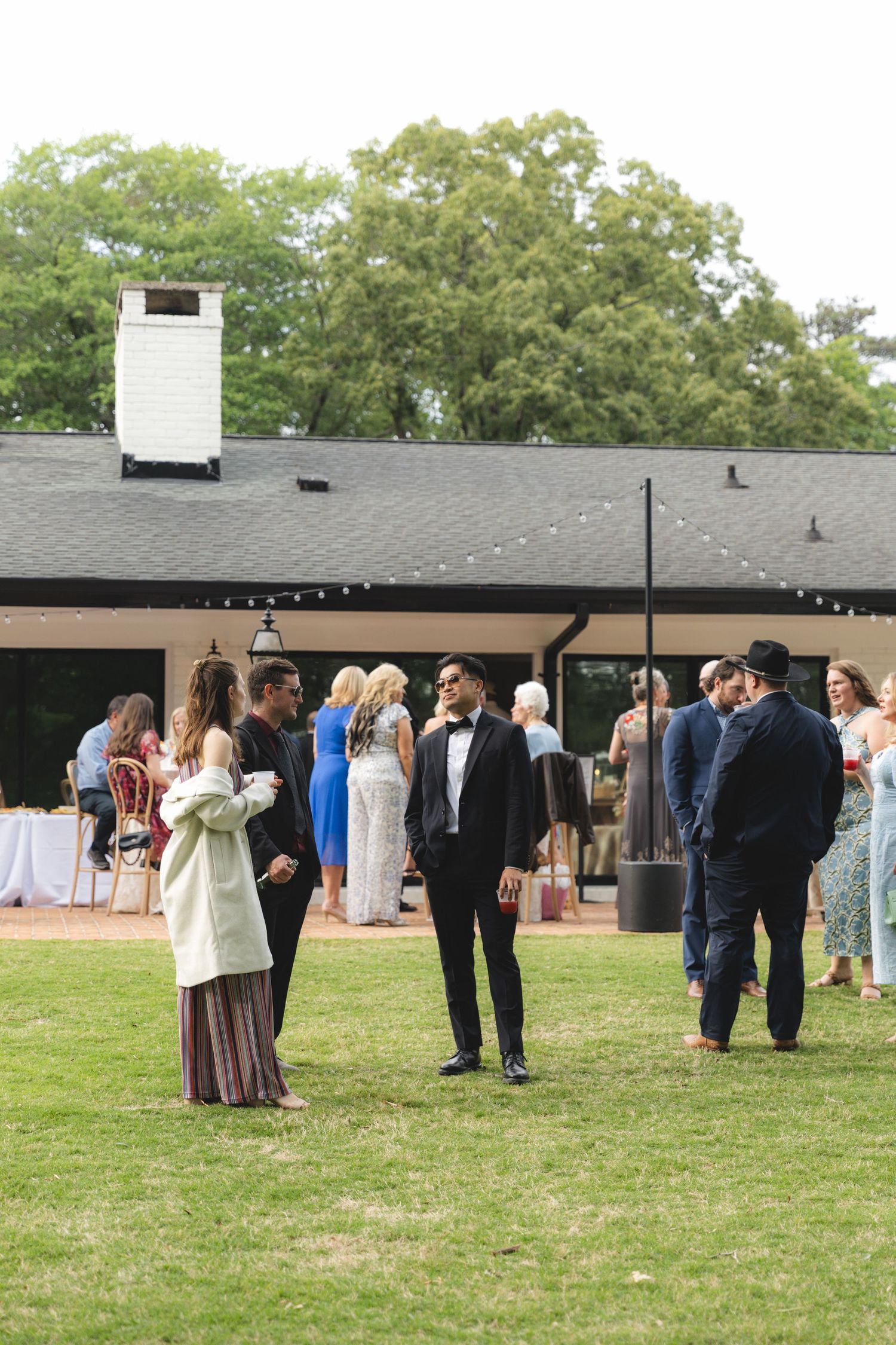 Guests mingle on green lawn outside elegant black venue building with string lights during outdoor wedding reception.