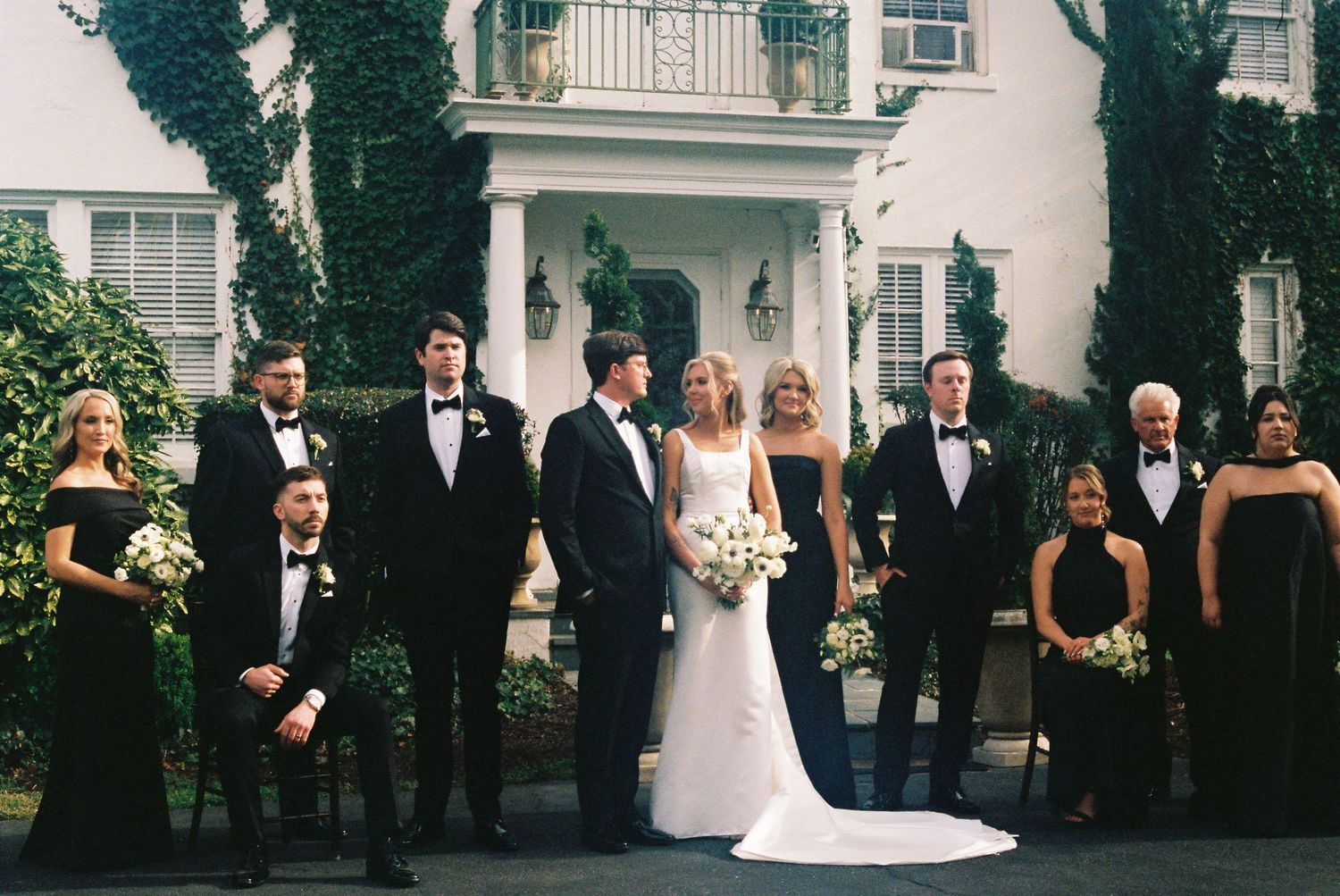 Wedding party in formal attire poses outside elegant white mansion, bride in white gown holding bouquet.