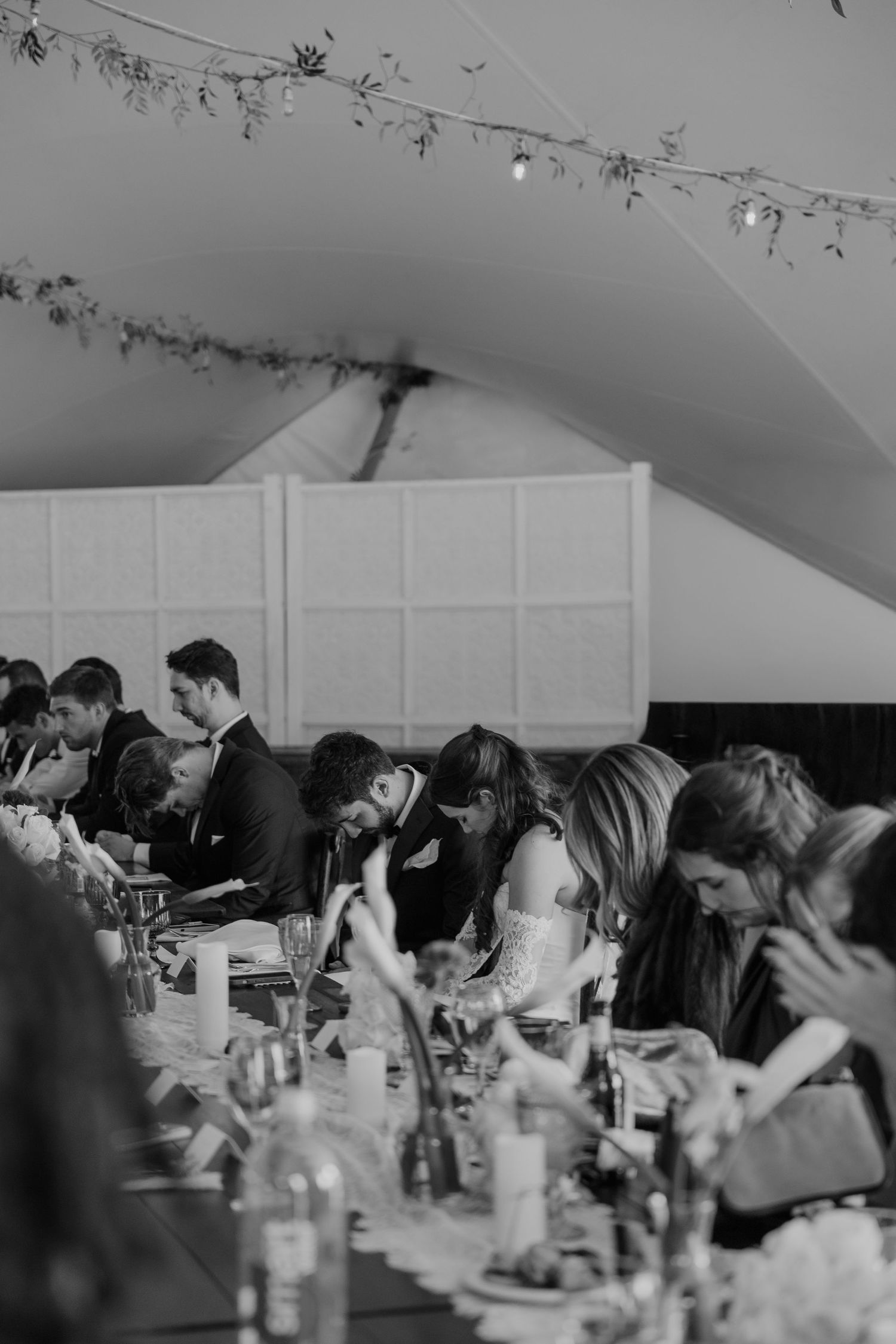 Black and white photo of wedding guests seated at a long reception table under a tent with string lights.