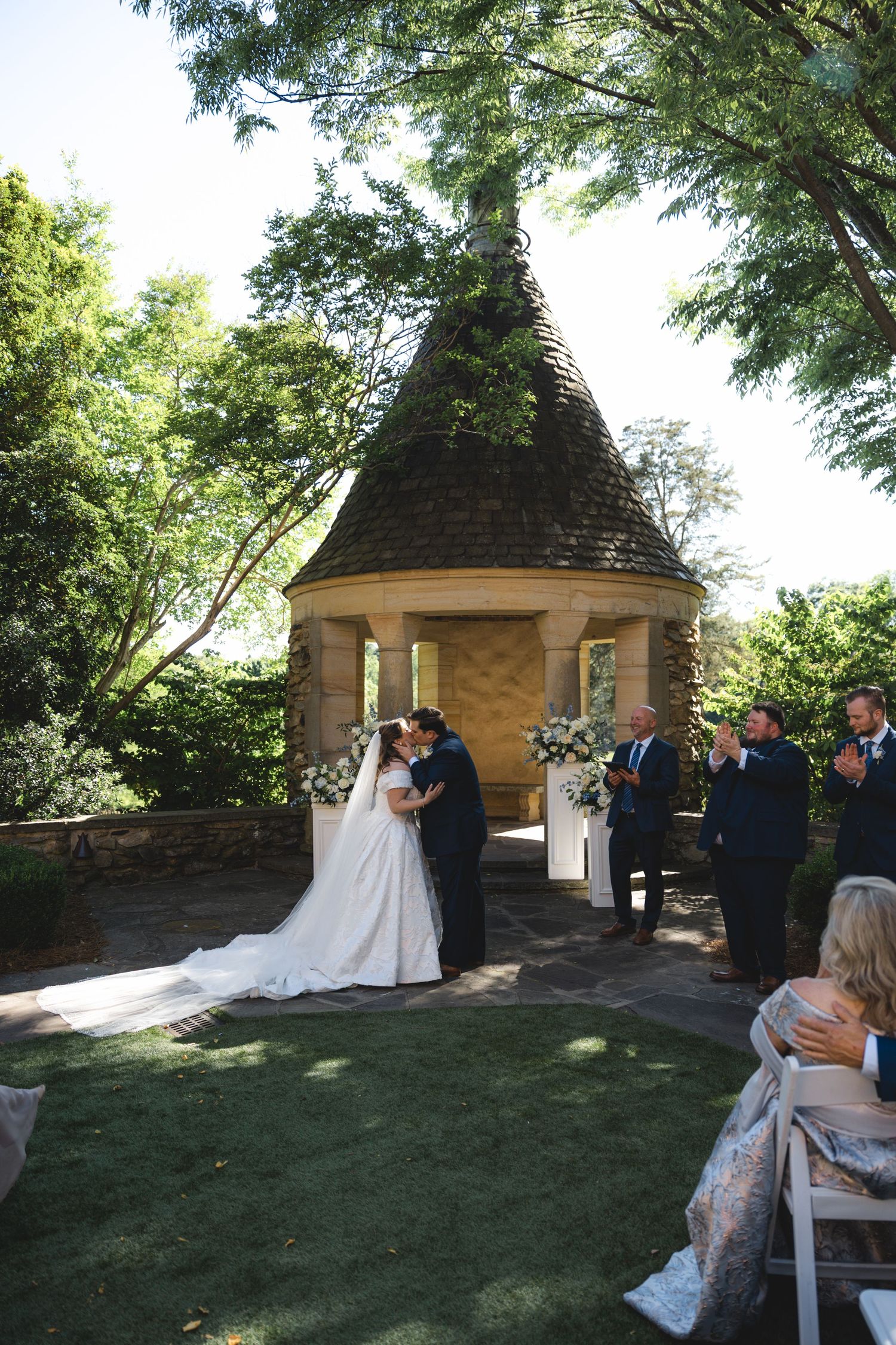 Bride and groom share first kiss at outdoor garden wedding ceremony beside stone gazebo with floral arrangements.
