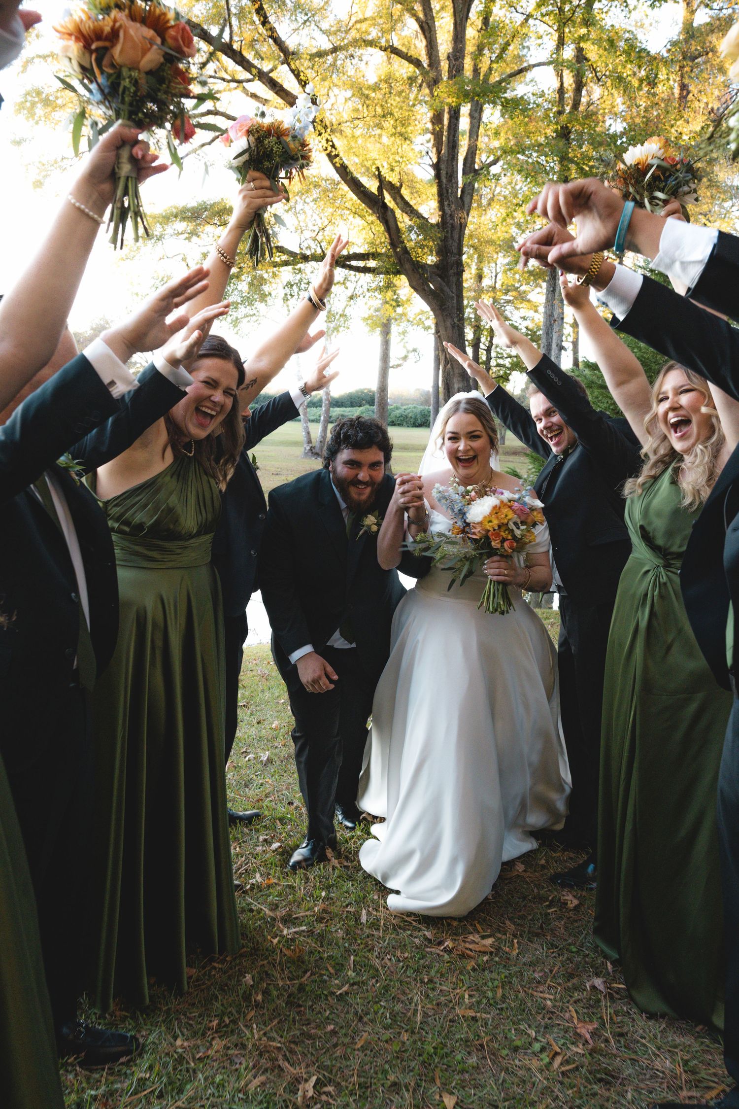Wedding party in green dresses celebrating outdoors with raised hands and bouquets.