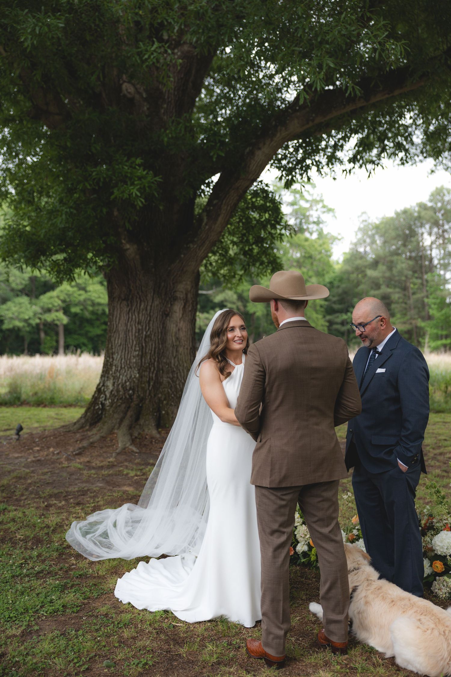 Bride and groom exchanging vows under large oak tree with officiant and dog at ceremony.