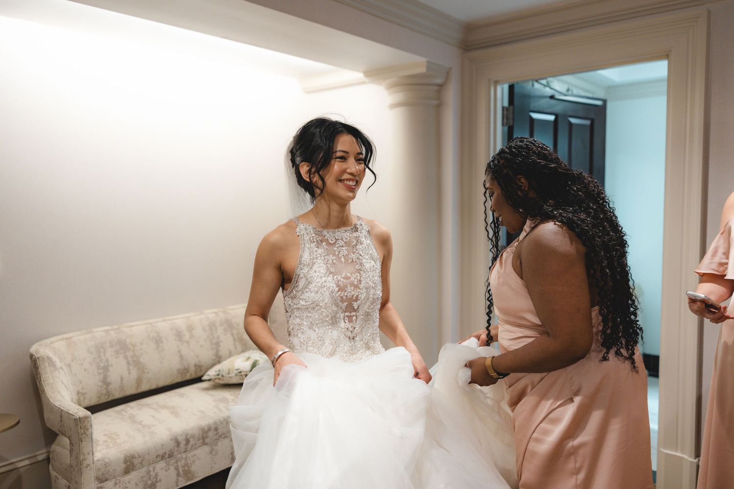 Joyful bride in beaded halter wedding gown laughing with bridesmaid in blush pink dress while getting ready.