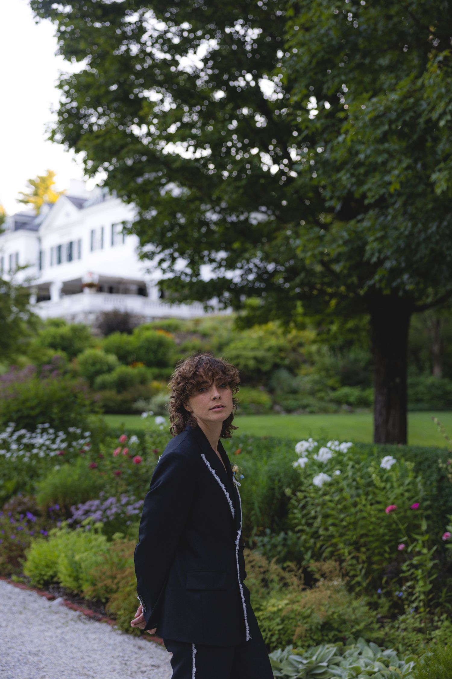 Person in black jacket standing on garden path with lush greenery and white house in background.